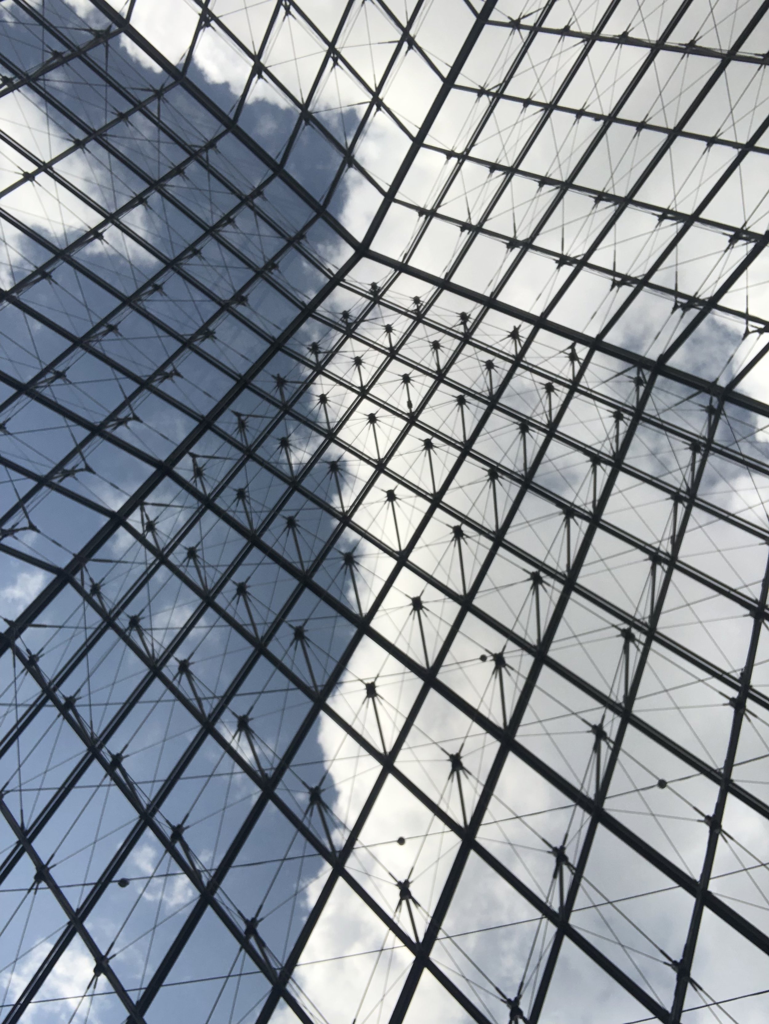 Looking up at the glass and metal lattice ceiling of the Louvre in Paris, France with a view of the cloudy sky.