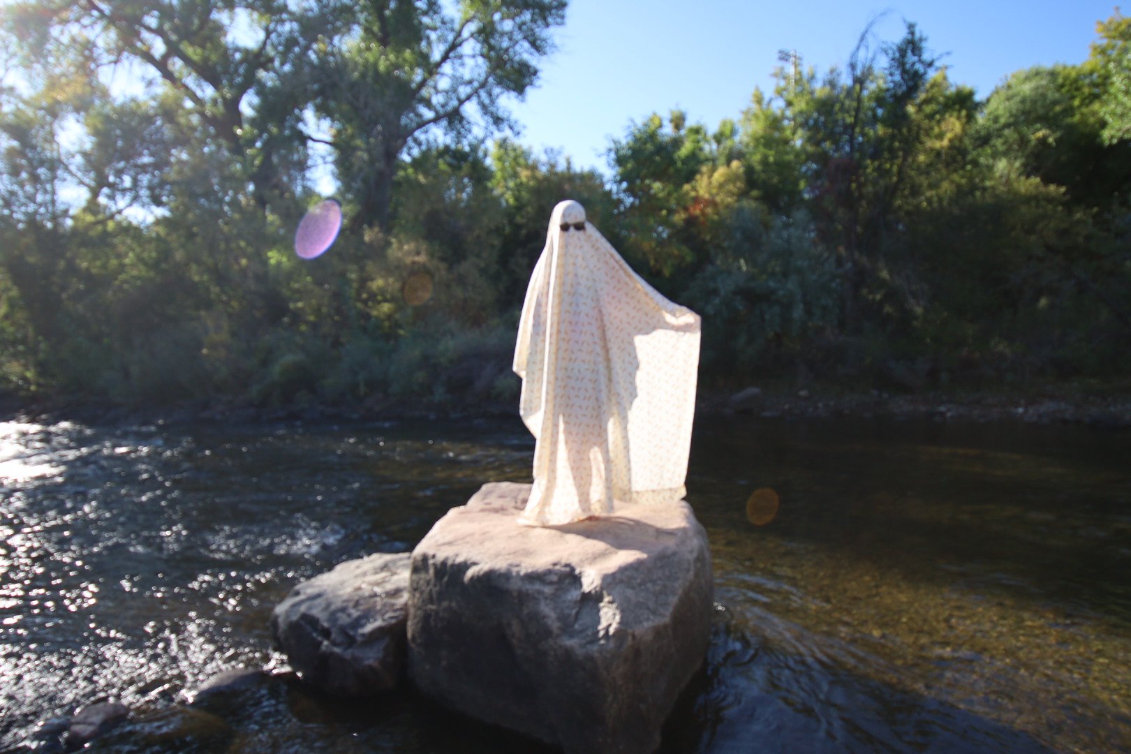 A cloth draped over a person dressing as a ghost, standing on a large rock in a river with trees and blue sky in the background.