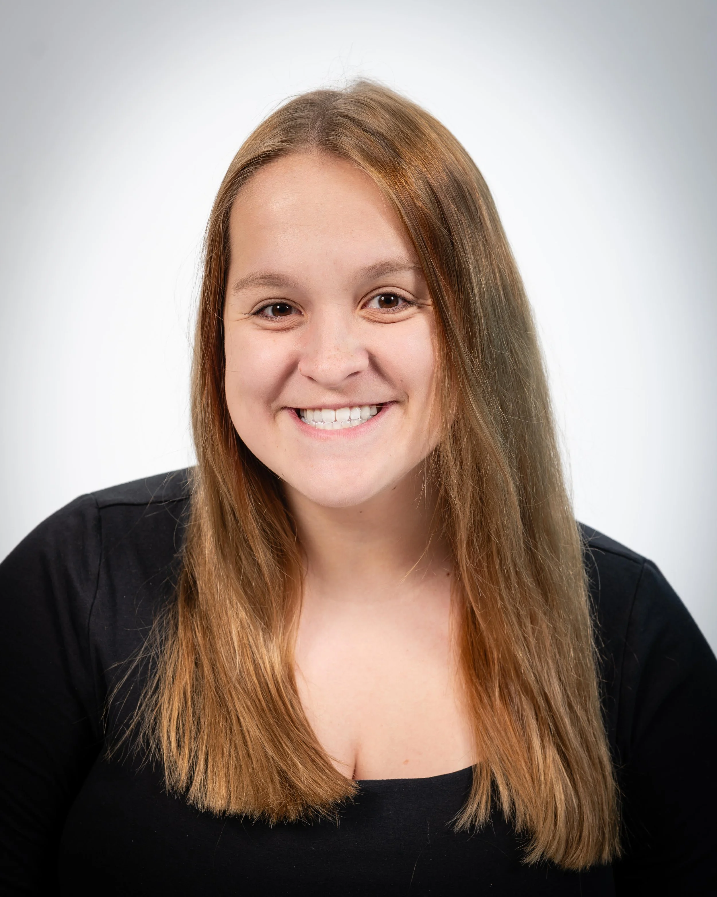 A smiling young woman with shoulder-length light brown hair, wearing a black top, against a plain white background.