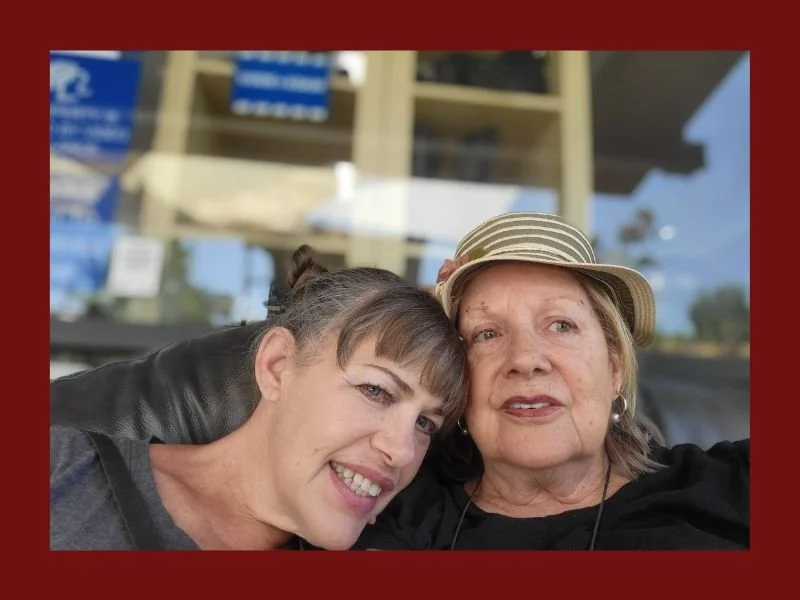 Two women, one younger and one older, sitting close together and smiling at the camera, with a store window in the background.