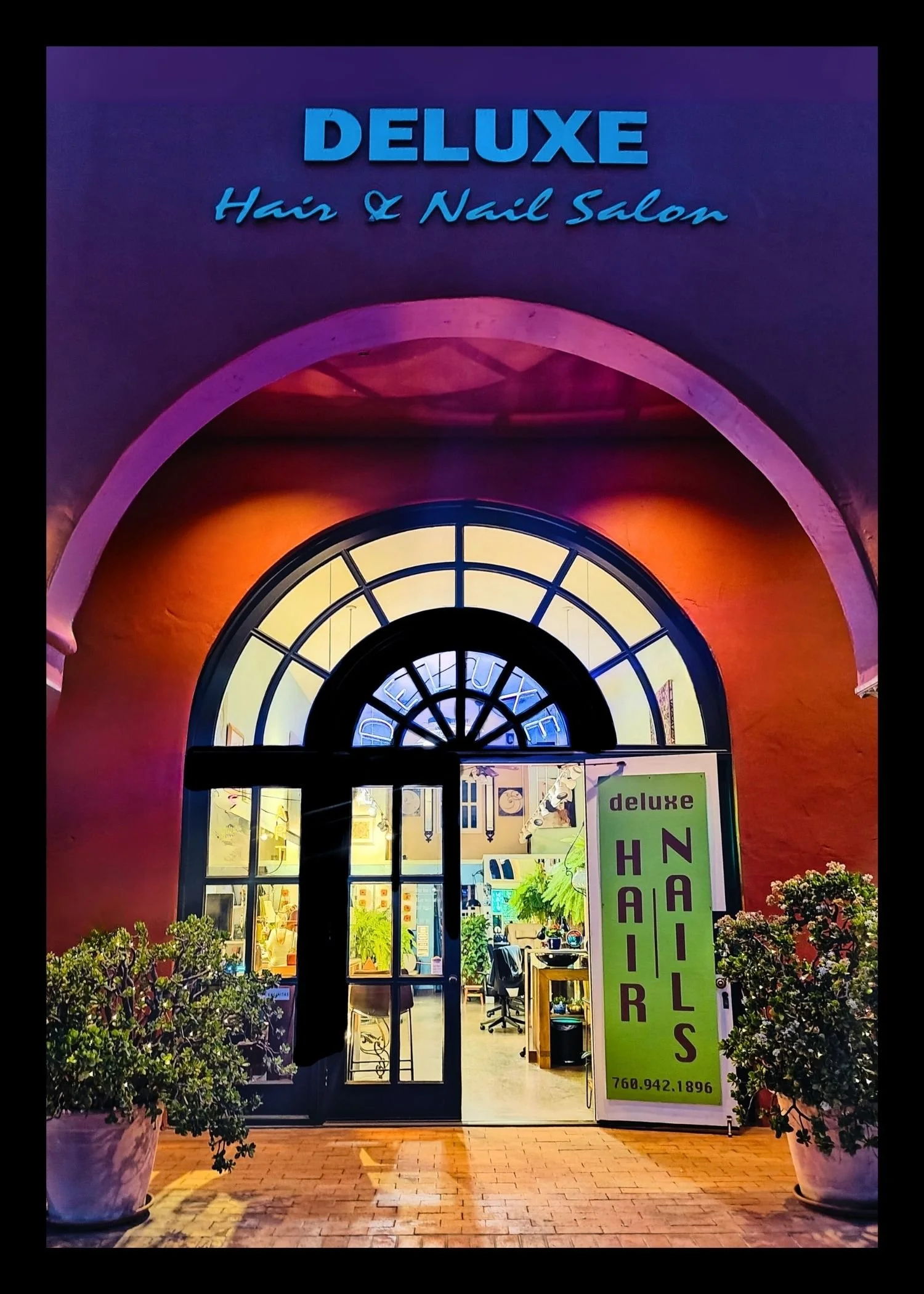 Exterior view of Deluxe Hair & Nail Salon with a brightly lit sign, large arched window, and potted plants outside at night.