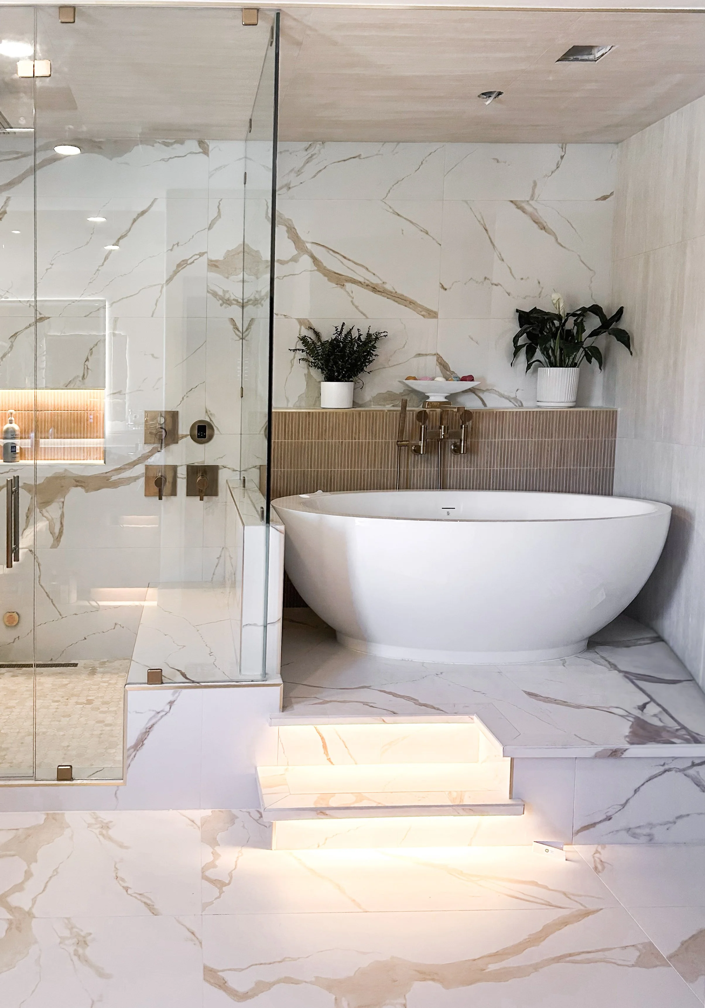 Luxurious spa-like bathroom with a freestanding bathtub, a wooden vanity with black countertops, hanging pendant lights, bird of paradise, a large window, and a floor length vanity mirrors.