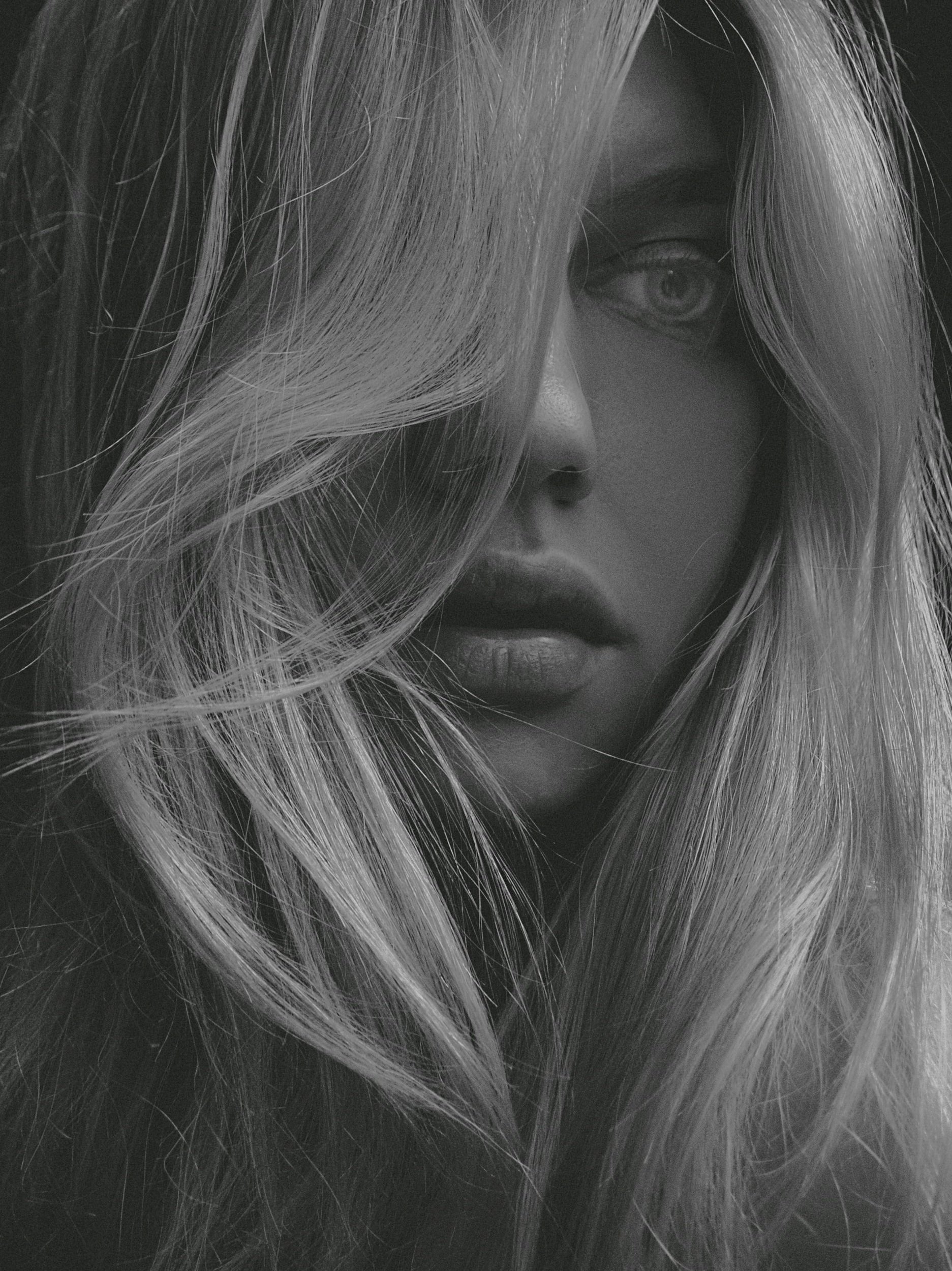 Close-up of a woman with long, blonde hair covering part of her face, gray eyes, and lips visible, black and white photo.