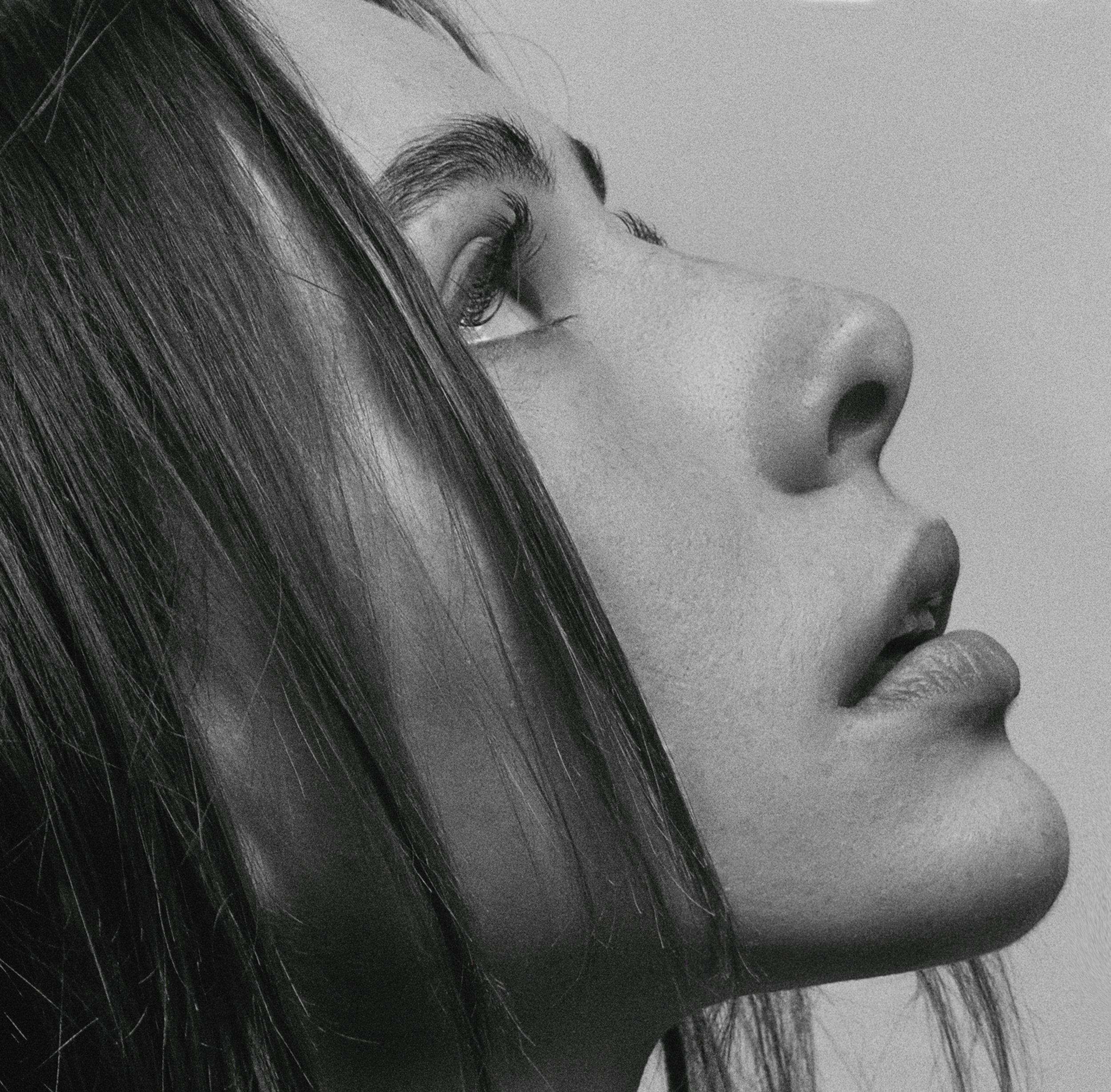 Close-up black-and-white photo of a woman's face, focusing on her eye with long eyelashes, eyebrows, and part of her nose and lips visible, with smooth skin and straight hair.