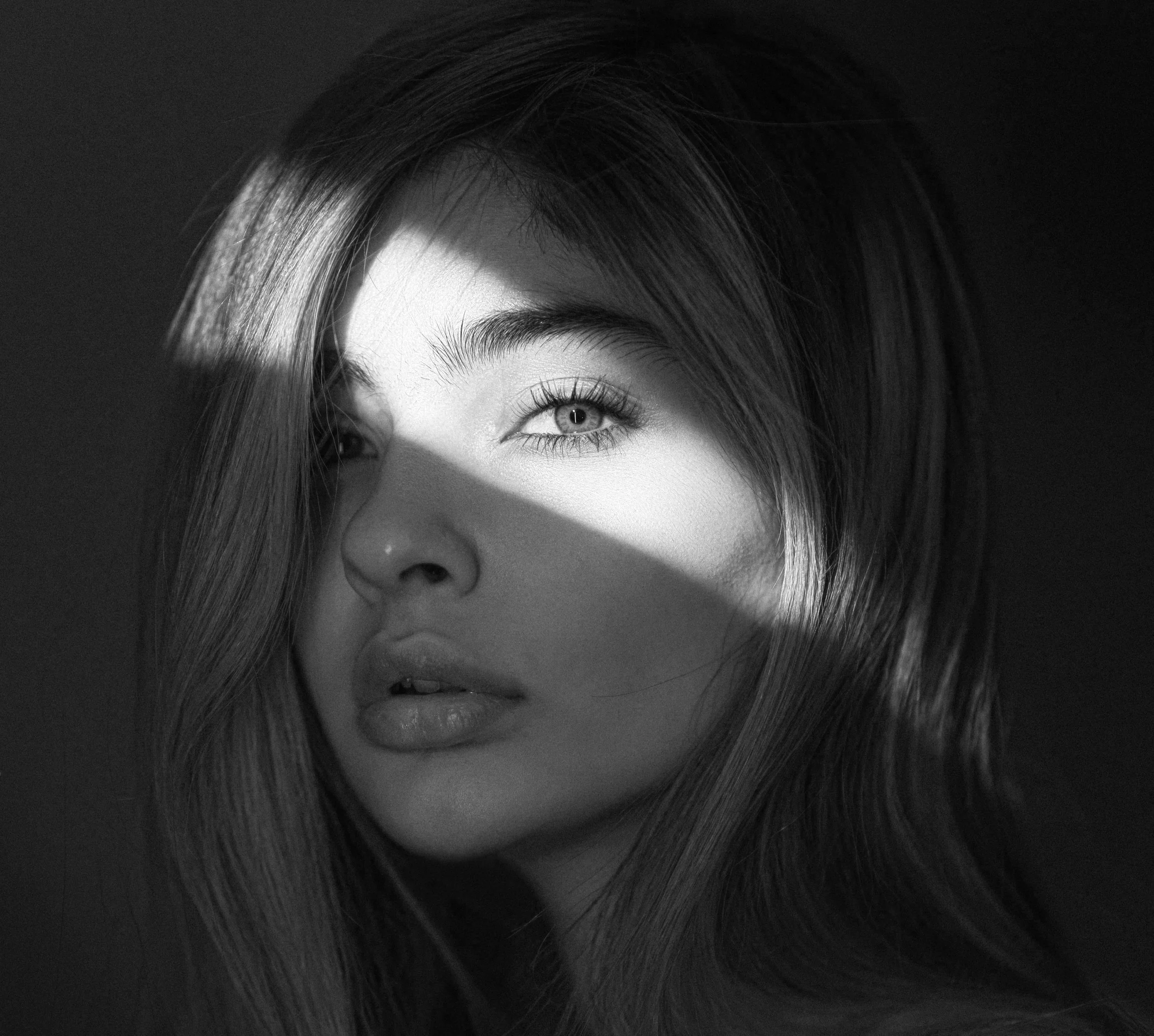 Close-up of a woman's face with light skin, focusing on her eye, eyebrow, and part of her forehead, with straight hair partially covering her face, shot in black and white.