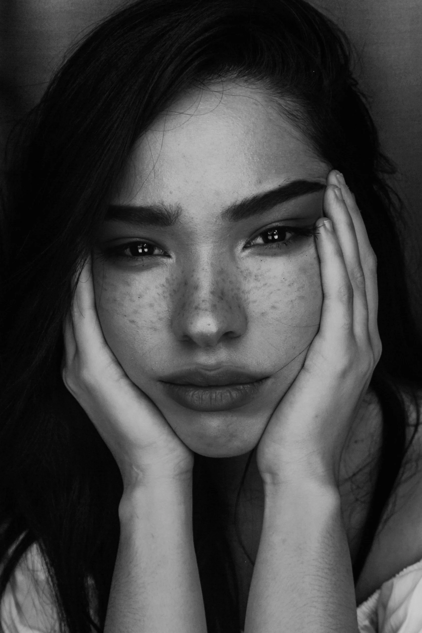 A close-up black and white photo of a young woman resting her face in her hands, with her fingers covering parts of her cheeks and forehead. She has dark hair, freckles, and expressive eyes.