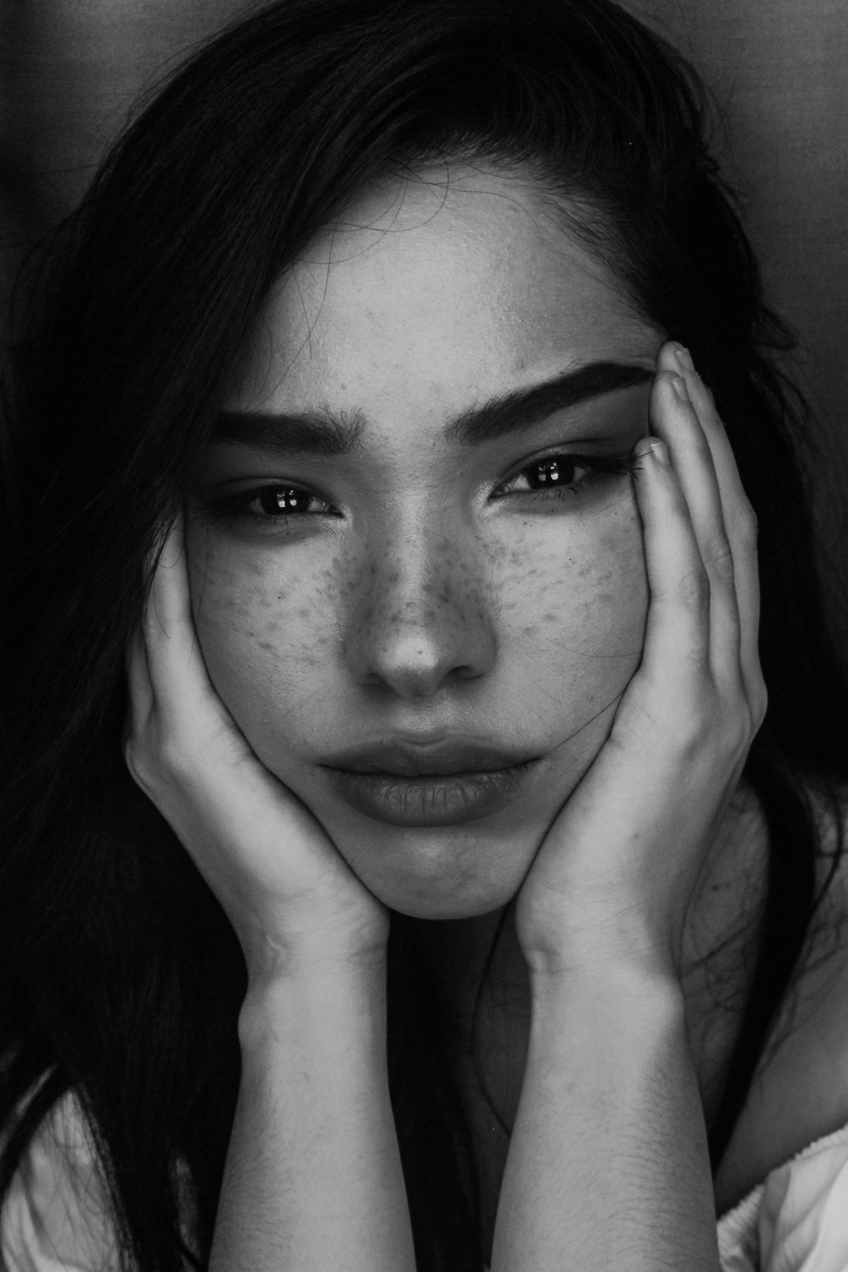 A black and white photo of a woman with long dark hair, facial freckles, and a gentle expression, resting her head in her hands.