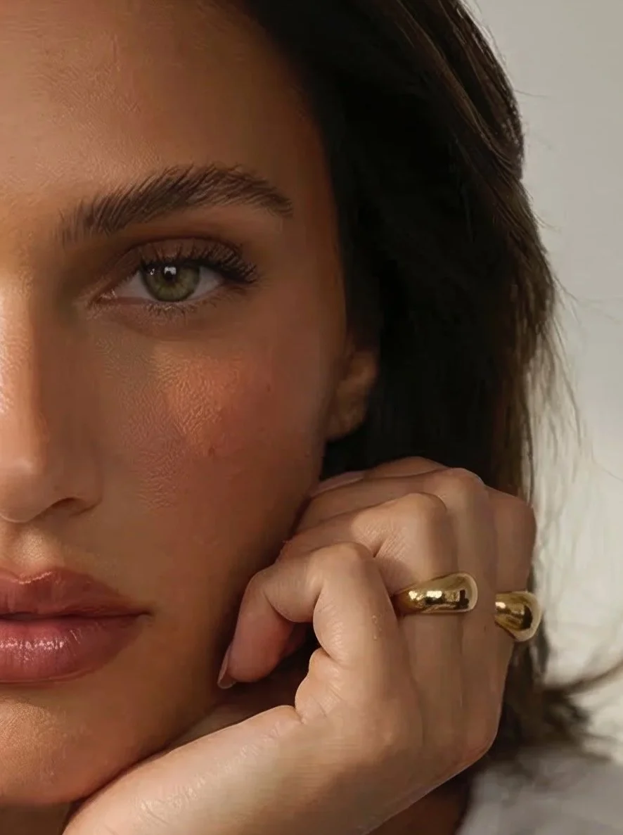 Close-up of a woman with light skin, green eyes, and dark hair, wearing a gold ring on her finger, resting her hand near her face.