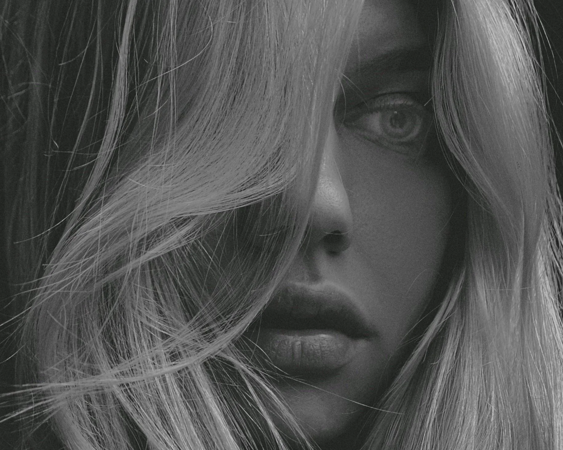 Close-up black and white photograph of a woman's face, partially obscured by long, blonde hair, with a focus on her eye and lips.