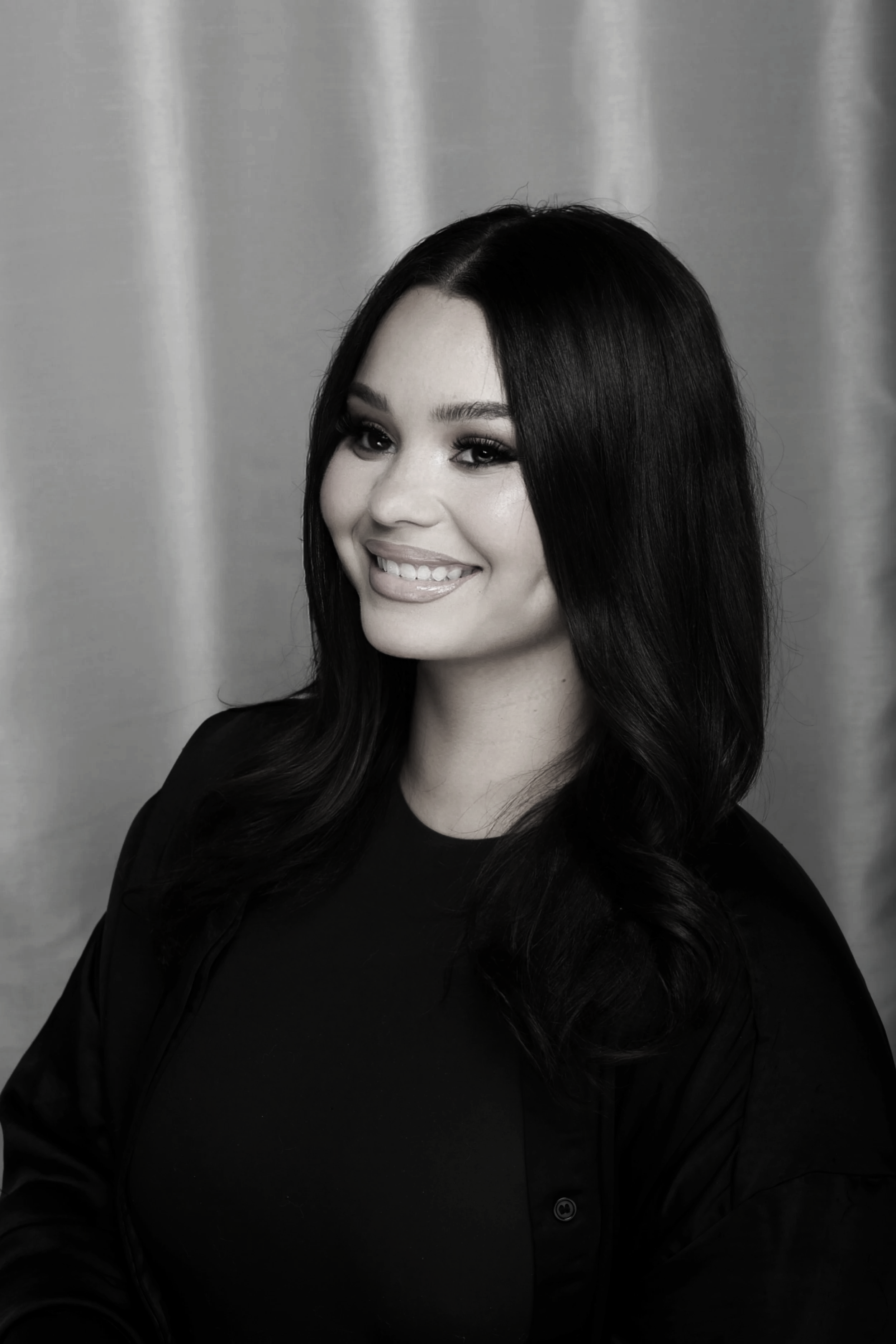 A woman with long dark hair and a bright smile, wearing a black top and blazer, posing in front of a neutral background.