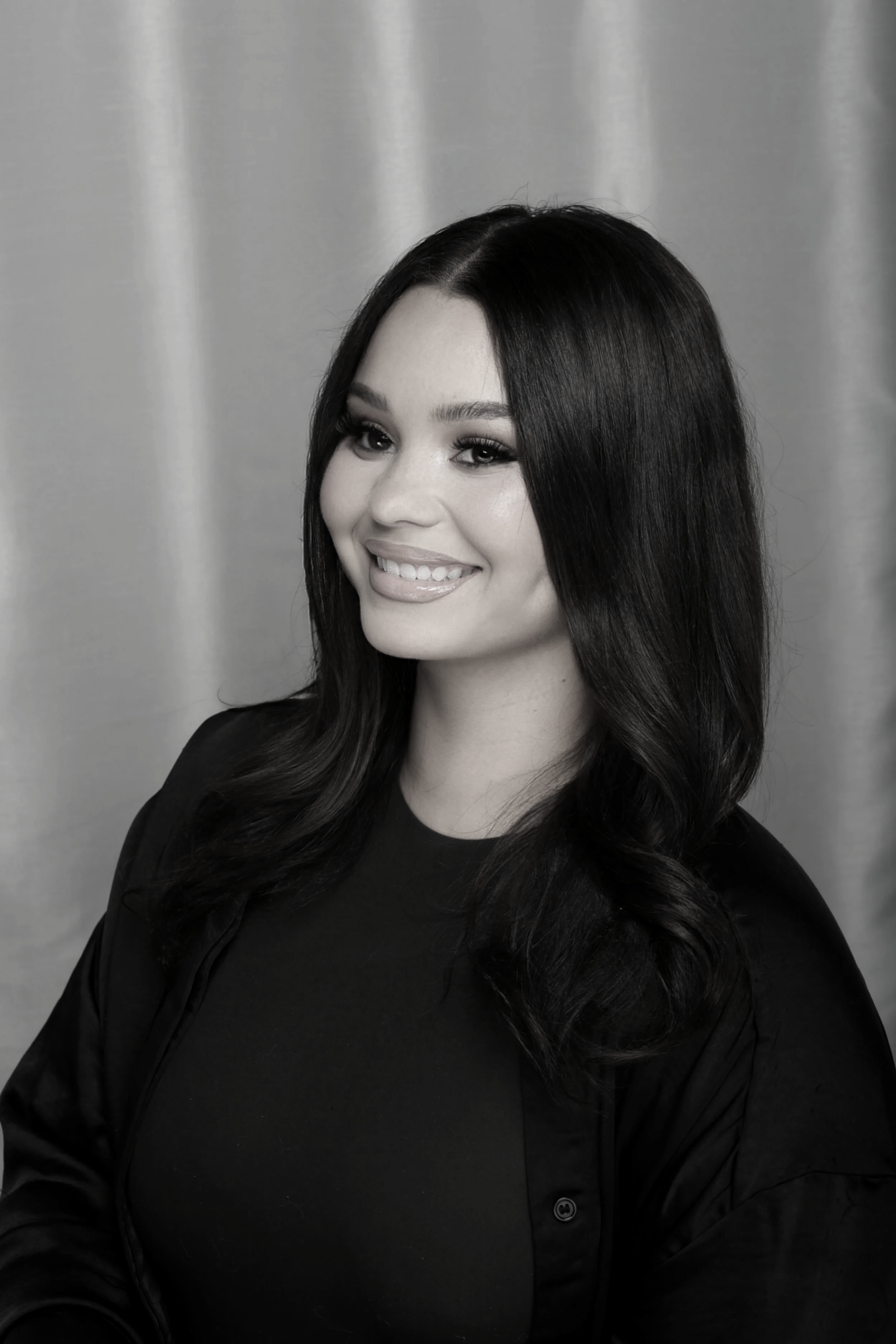 A woman with long, wavy dark hair, smiling and wearing a black top and jacket, standing against a plain background.