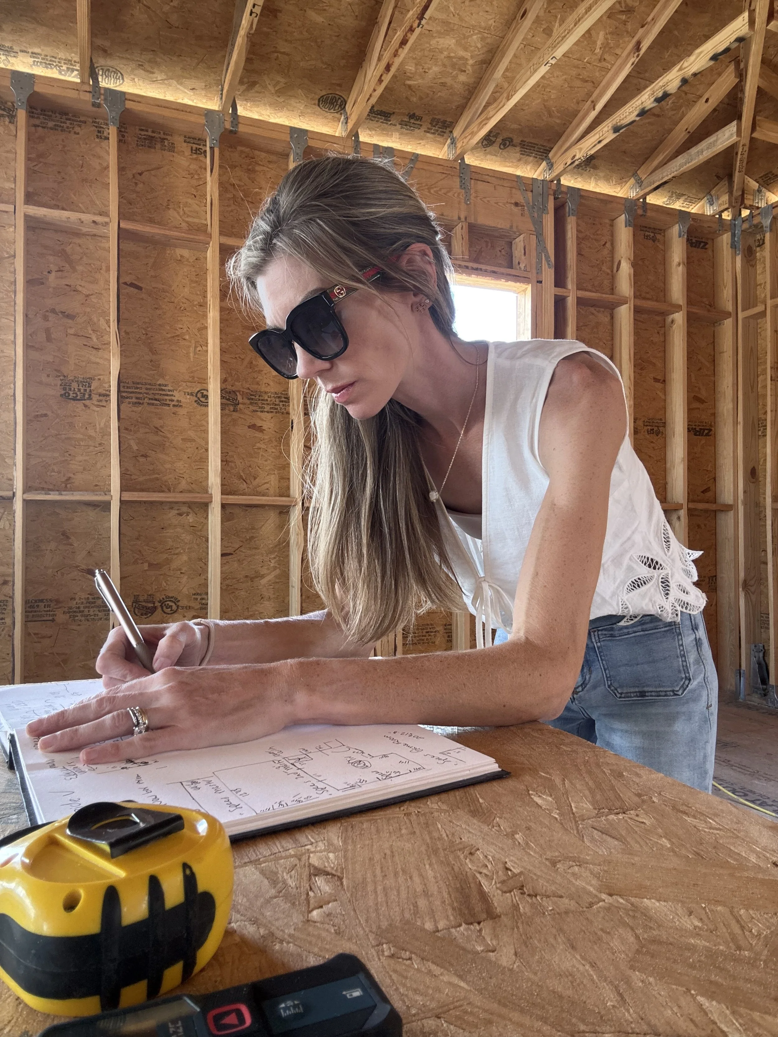 A woman with long blonde hair wearing sunglasses and a white sleeveless top writing in a notebook at a wooden worksite with unfinished walls and a window, with a yellow tape measure and digital device on the table.