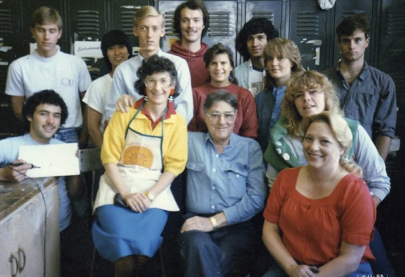 Lydia Ruyle and her art students. Group includes eleven people, including two women and nine young men, posing in a room with lockers in the background. They appear happy and are smiling for the photo.