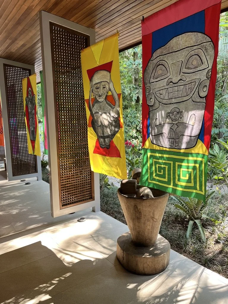 Colorful fabric art Goddess Banner hanging in Costa Rica with abstract human and face designs hanging in a garden-like space, with potted plants and sunlight casting shadows.