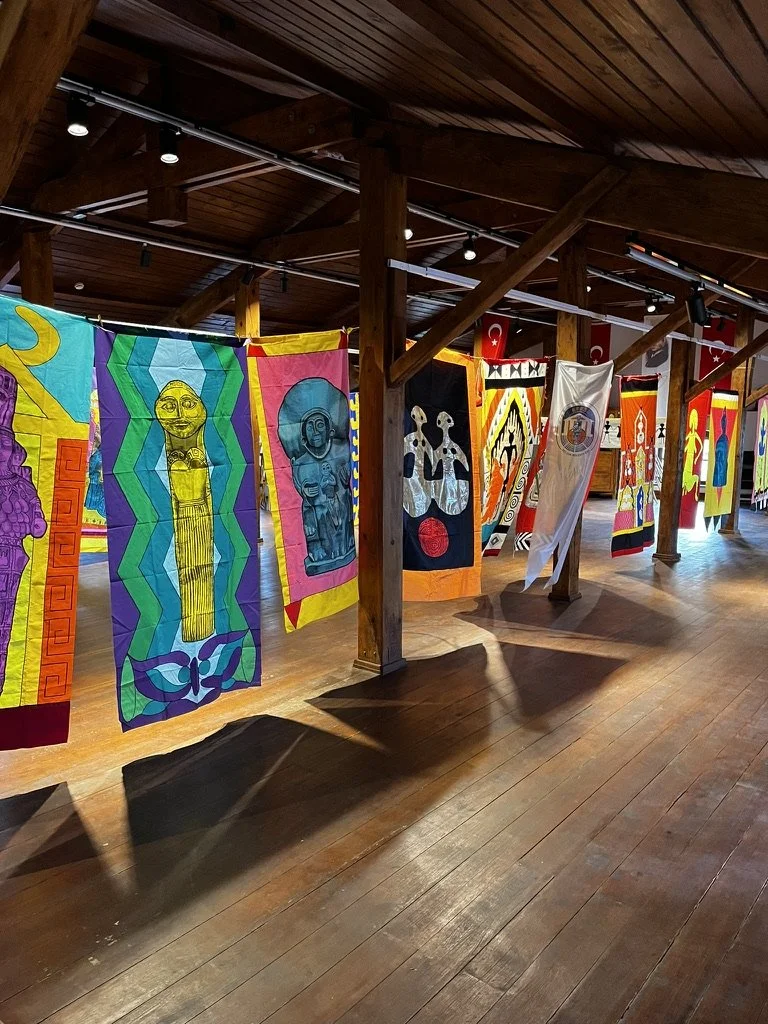 Colorful goddess banner artwork by Lydia Ruyle hanging in the Ephesus Museum on a line inside a wooden gallery, featuring ancient divine feminine images of Anatolia.
