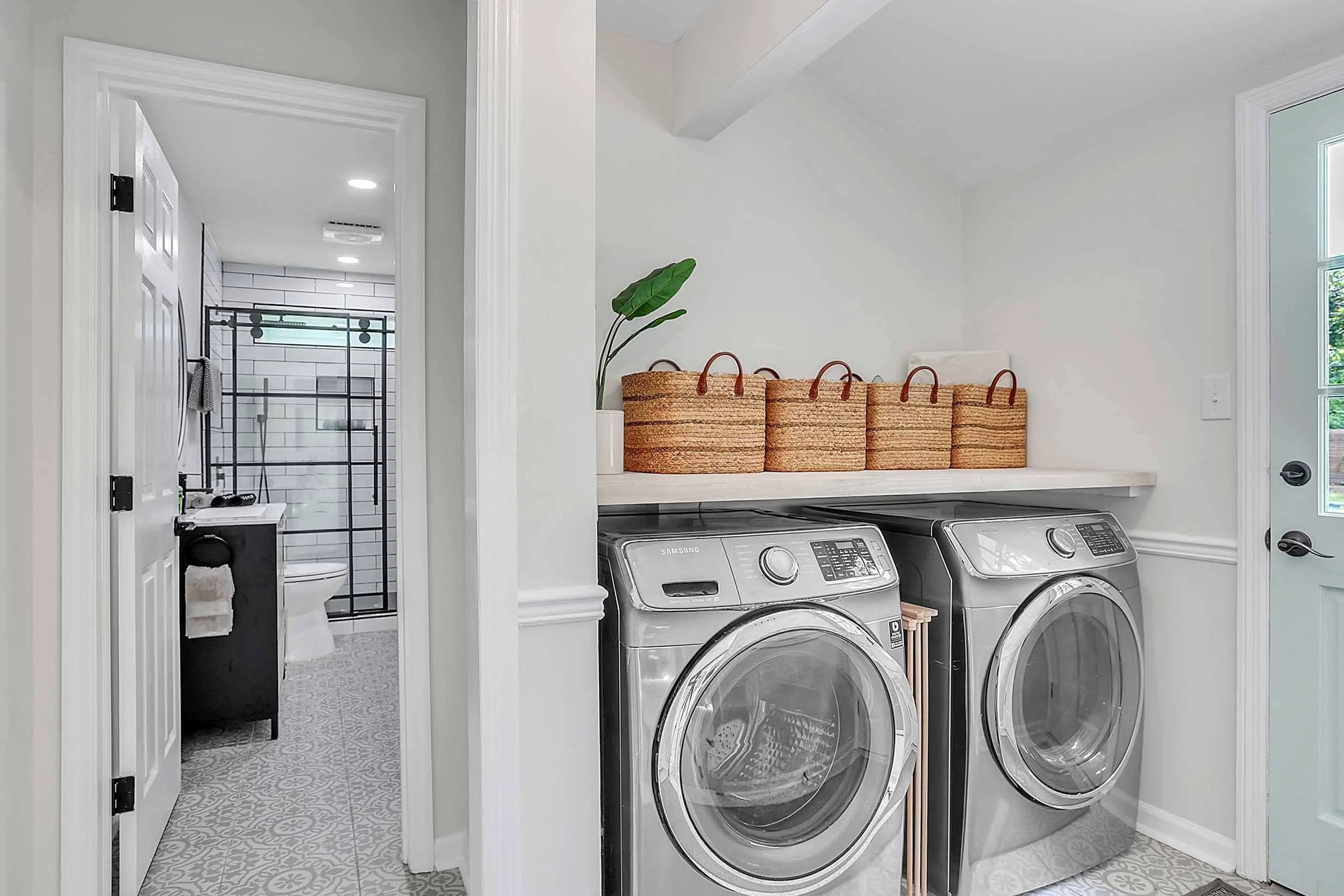 Laundry area with washer, dryer, bamboo rack, woven baskets on shelf, tropical plant, open door to modern bathroom, door to patio with grill, outdoor furniture, and heated pool in pet-friendly oasis