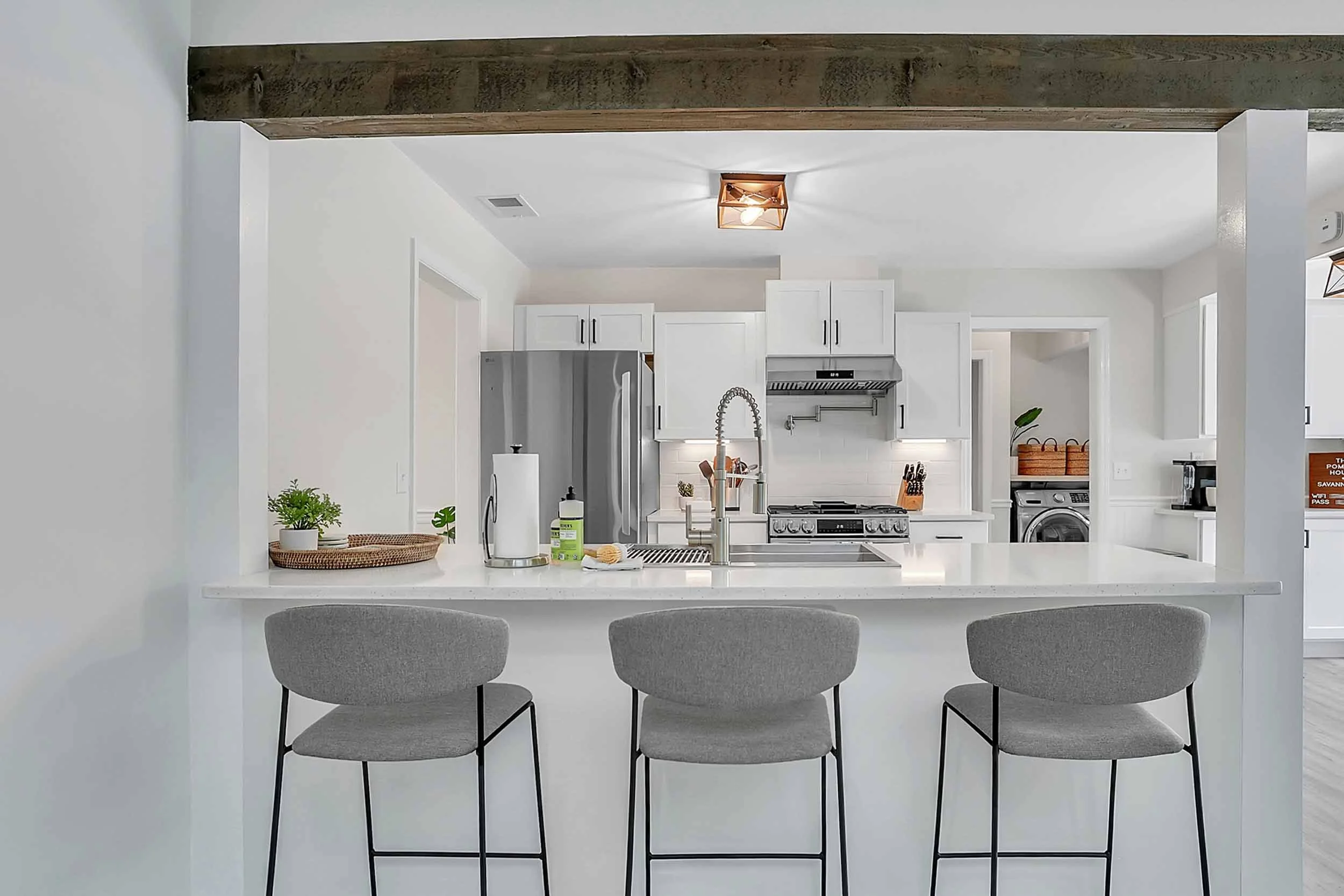 Bar seating at kitchen island with white quartz countertop, trio of Italian designer stools in taupe fabric with backrests and footrests, view to fully equipped chef’s kitchen with new appliances