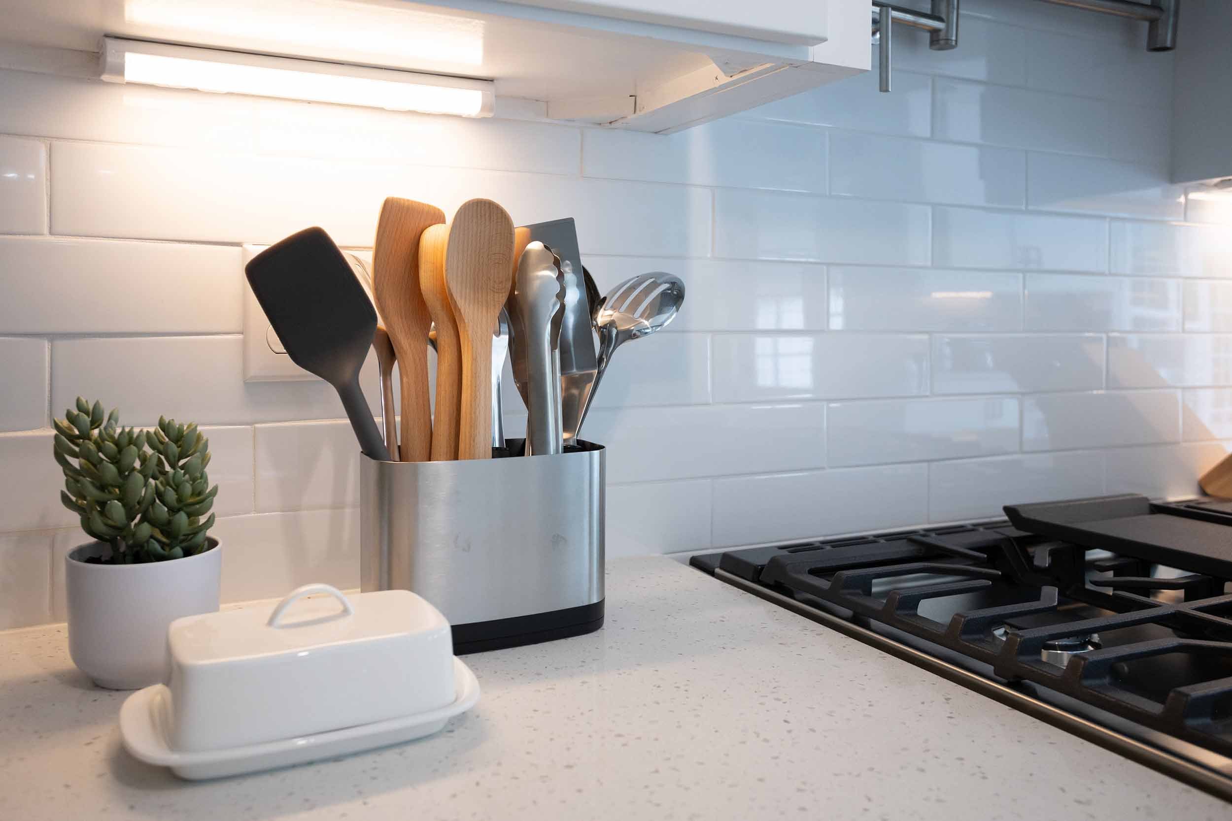 Closeup of gas range with white subway tile backsplash, variety of kitchen tools inside stainless utensil holder, and wide porcelain butter dish on quartz countertop, below warm under-cabinet lighting