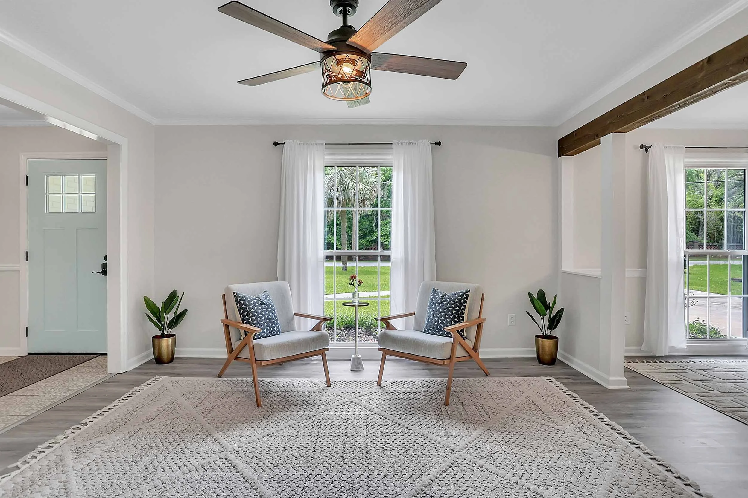 View of sitting room from living space showing spacious area rug, two lounge chairs, side table, tropical plants, large window overlooking yard with palm trees on Wilmington Island in Savannah, GA