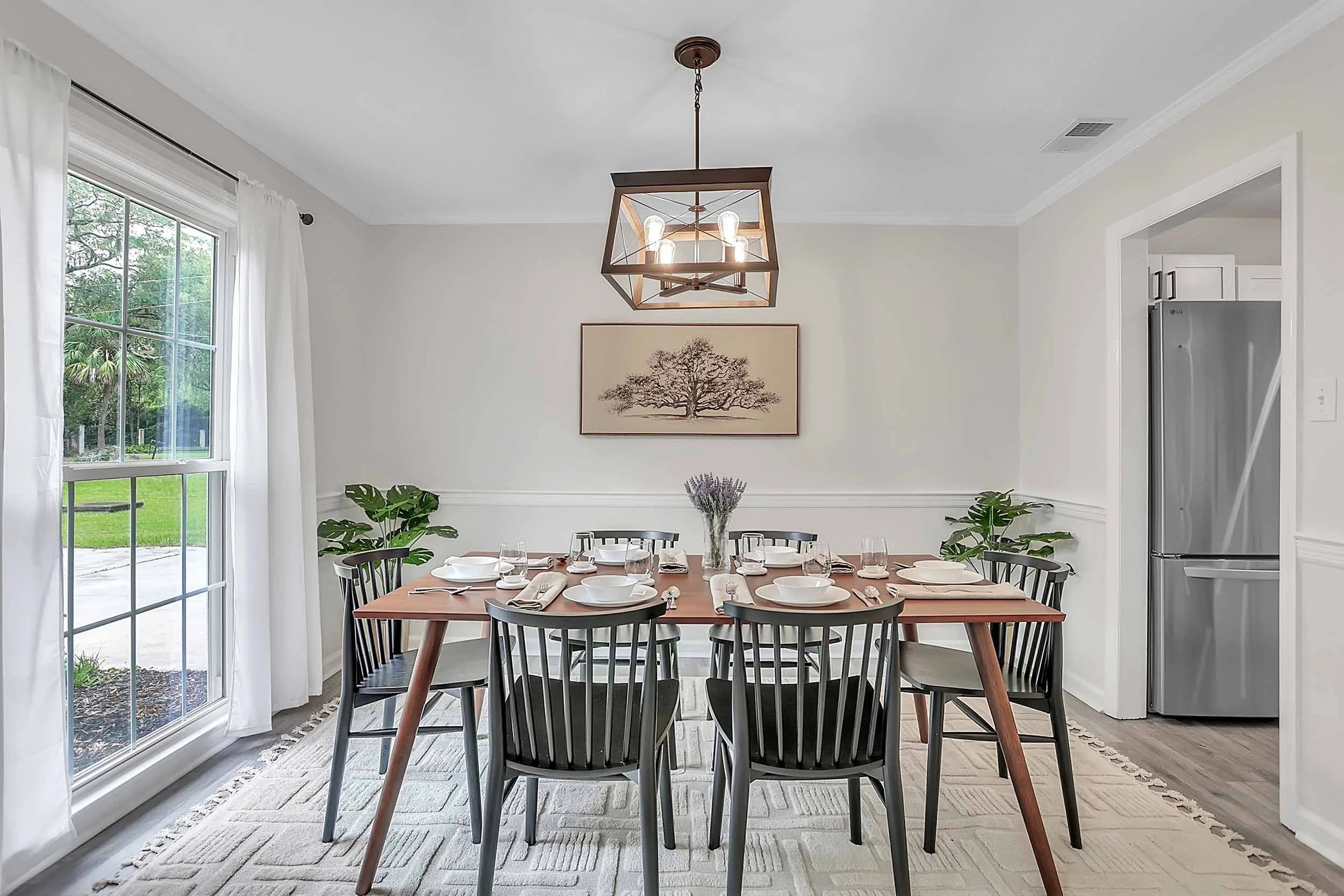 Dining room with large mid-century modern wood table set for meals, black Scandinavian chairs, southern oak art inspired by Savannah, rustic pendant light, oversized window with white sheer curtains