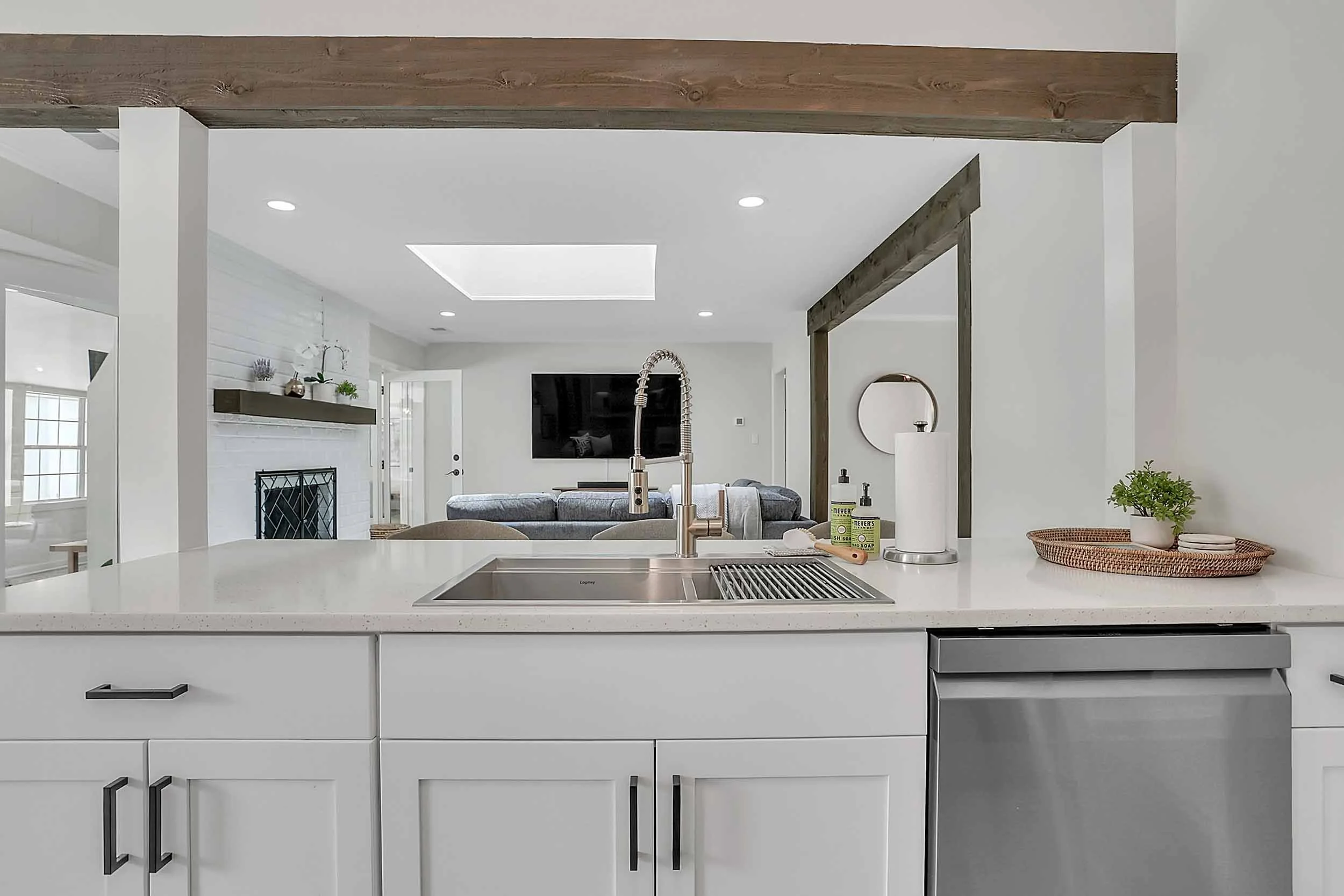 Kitchen island with sink, pull-down faucet, white shaker cabinets, quartz counters, stainless dishwasher, with view into living room of exposed wood beams, skylight, wall-mounted 77” TV with soundbar