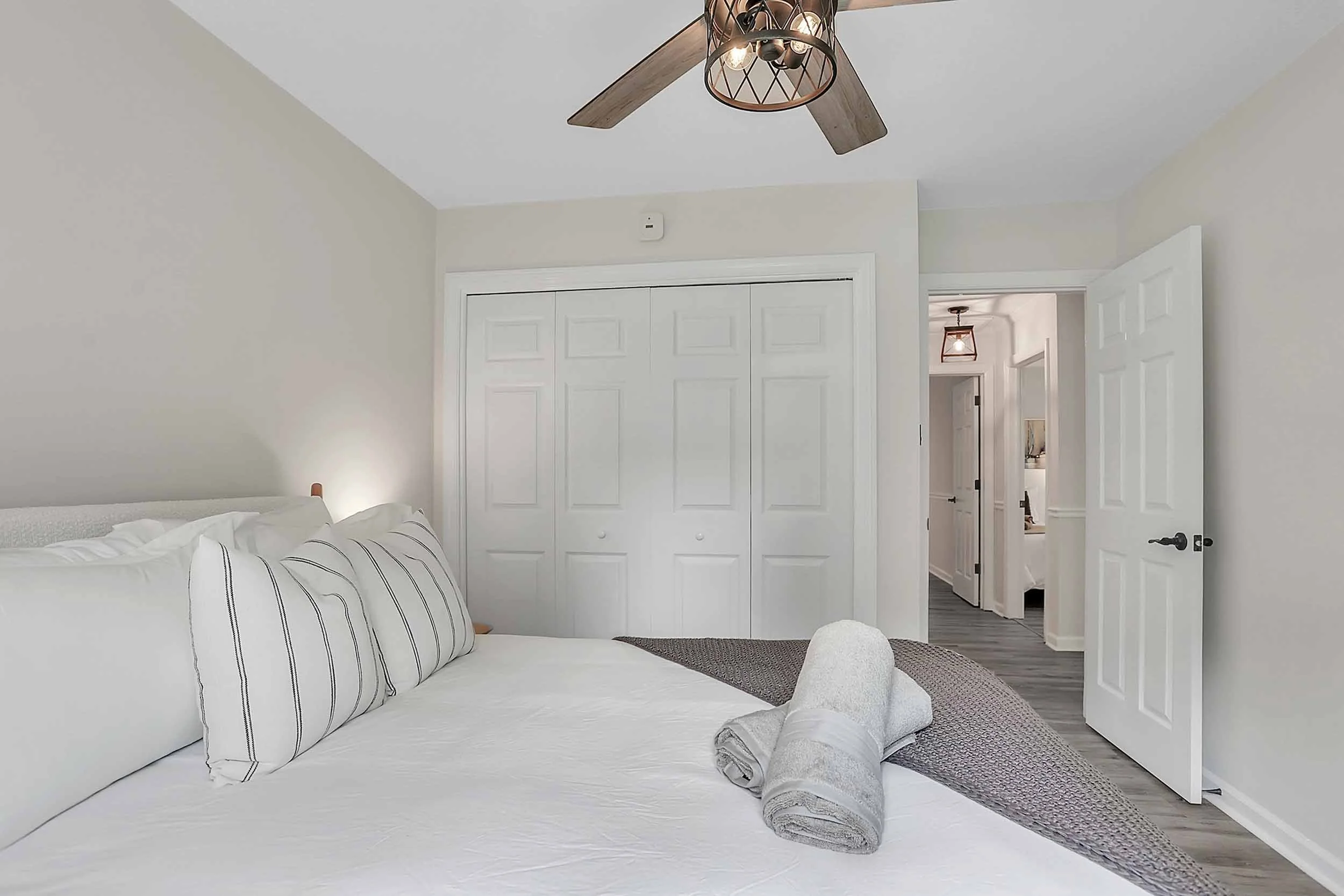 Alternate view of queen bedroom at Pomelo House Savannah vacation rental showing plush bed with striped pillows and knit throw, large bi-fold closet, ceiling fan, and view to hallway with bathroom