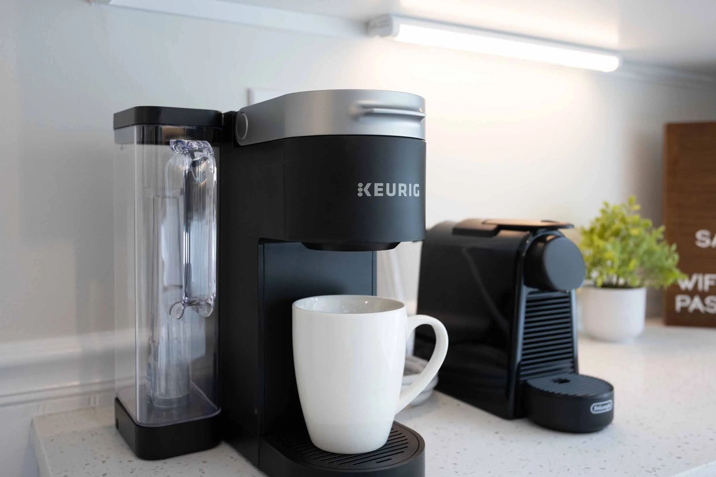 Coffee station at Pomelo House vacation rental home in Savannah, GA with Keurig coffee maker, porcelain mug, Nespresso machine, WiFi sign, white quartz countertop, warm under-cabinet lighting