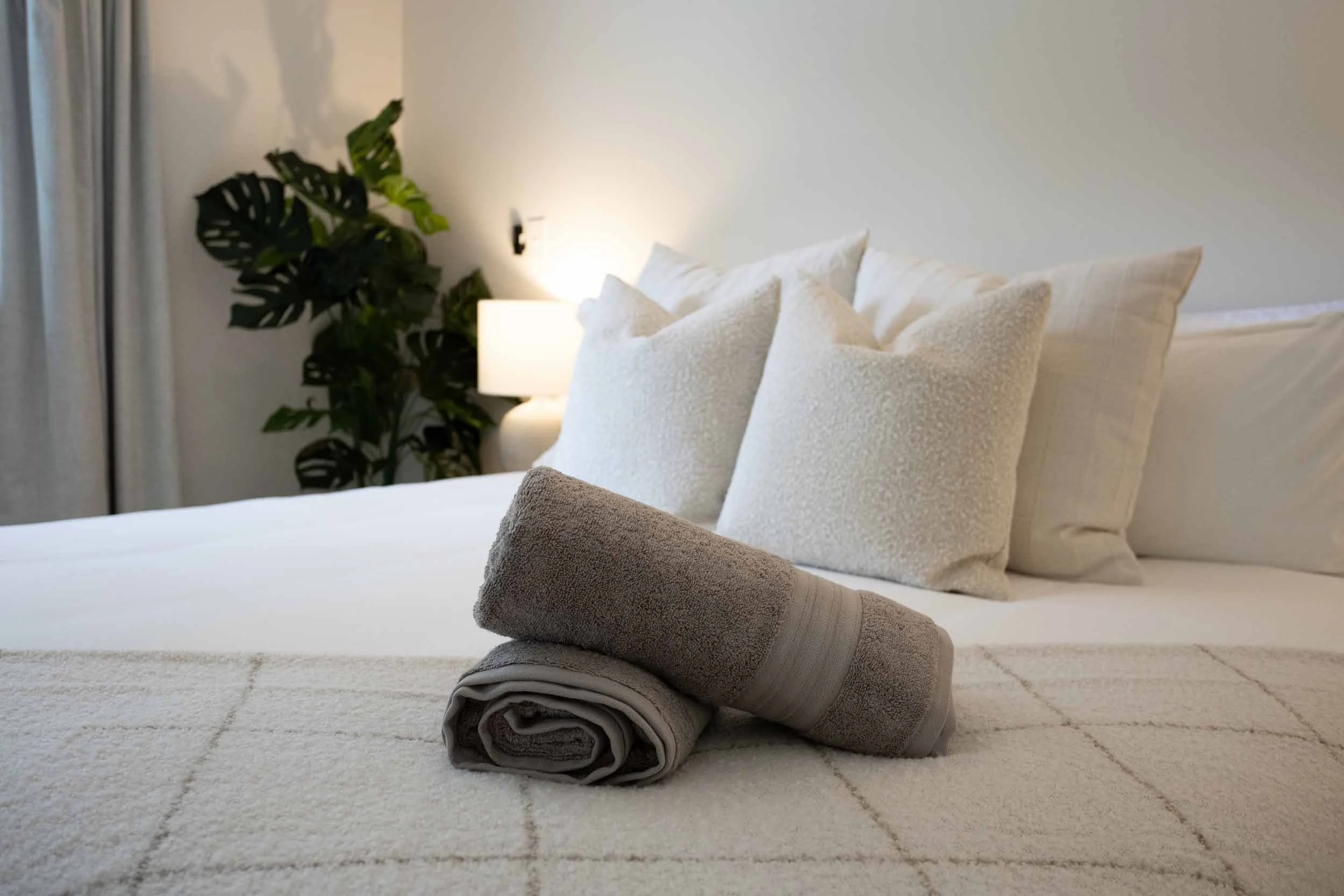 Closeup of king hybrid mattress, 100% Egyptian cotton linens, rolled cotton guest towels, textured throw blanket, layered white and beige pillows, warm lamp glow, blackout curtains, monstera plant