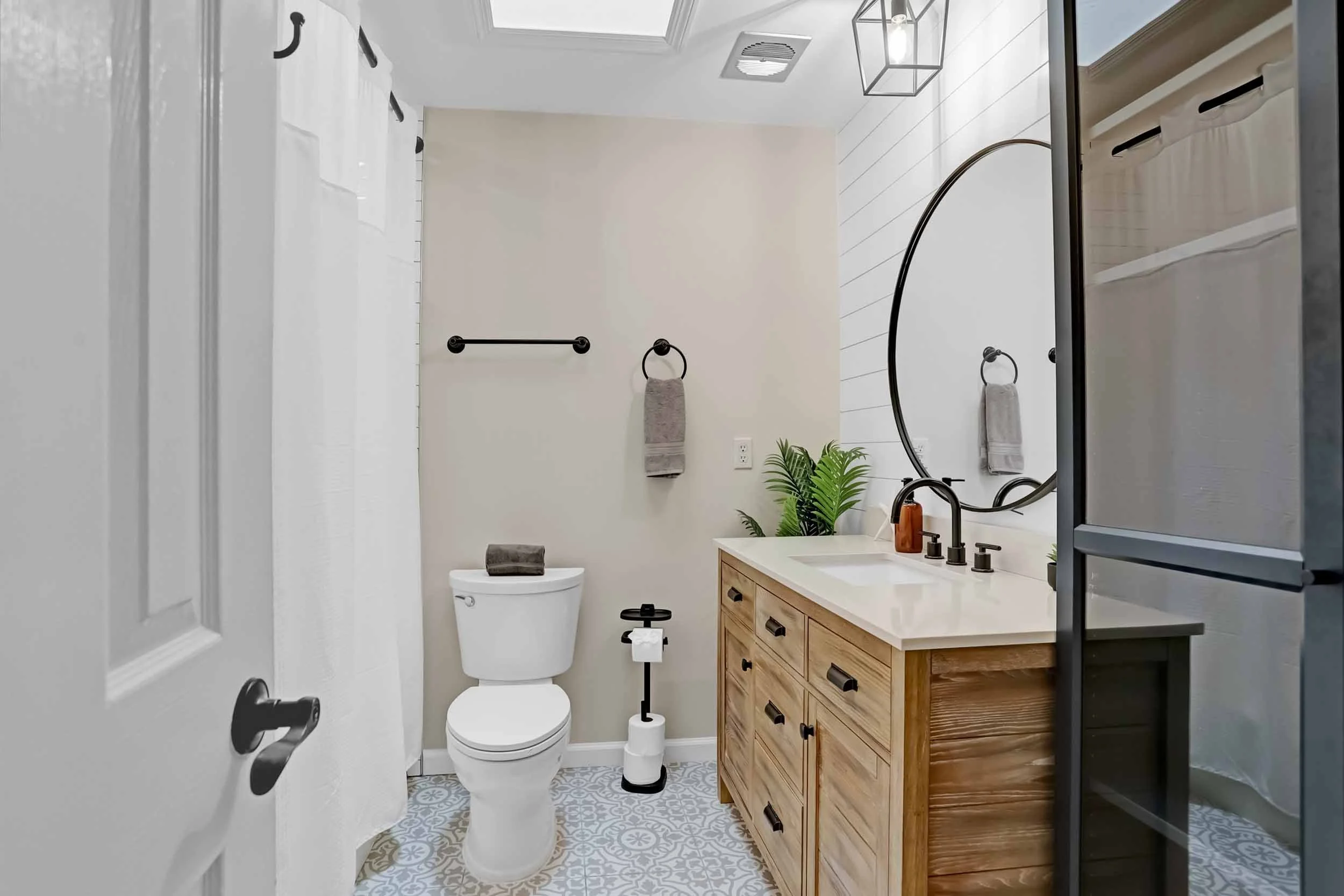 Beach-themed hall bathroom with tub shower, wood vanity with sand-colored quartz countertop and sink, round mirror, toilet, bookcase with glass doors, shiplap wall, palm tree, succulent, and starfish