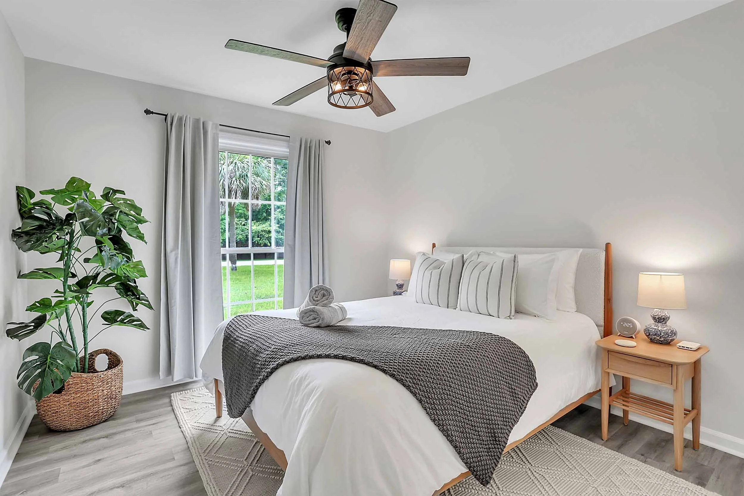 Queen bedroom with premium hybrid mattress, blackout curtains, Egyptian cotton linens, pillows, towels, throw blanket, side tables, lamps, oversized window looking out to street lined with palm trees