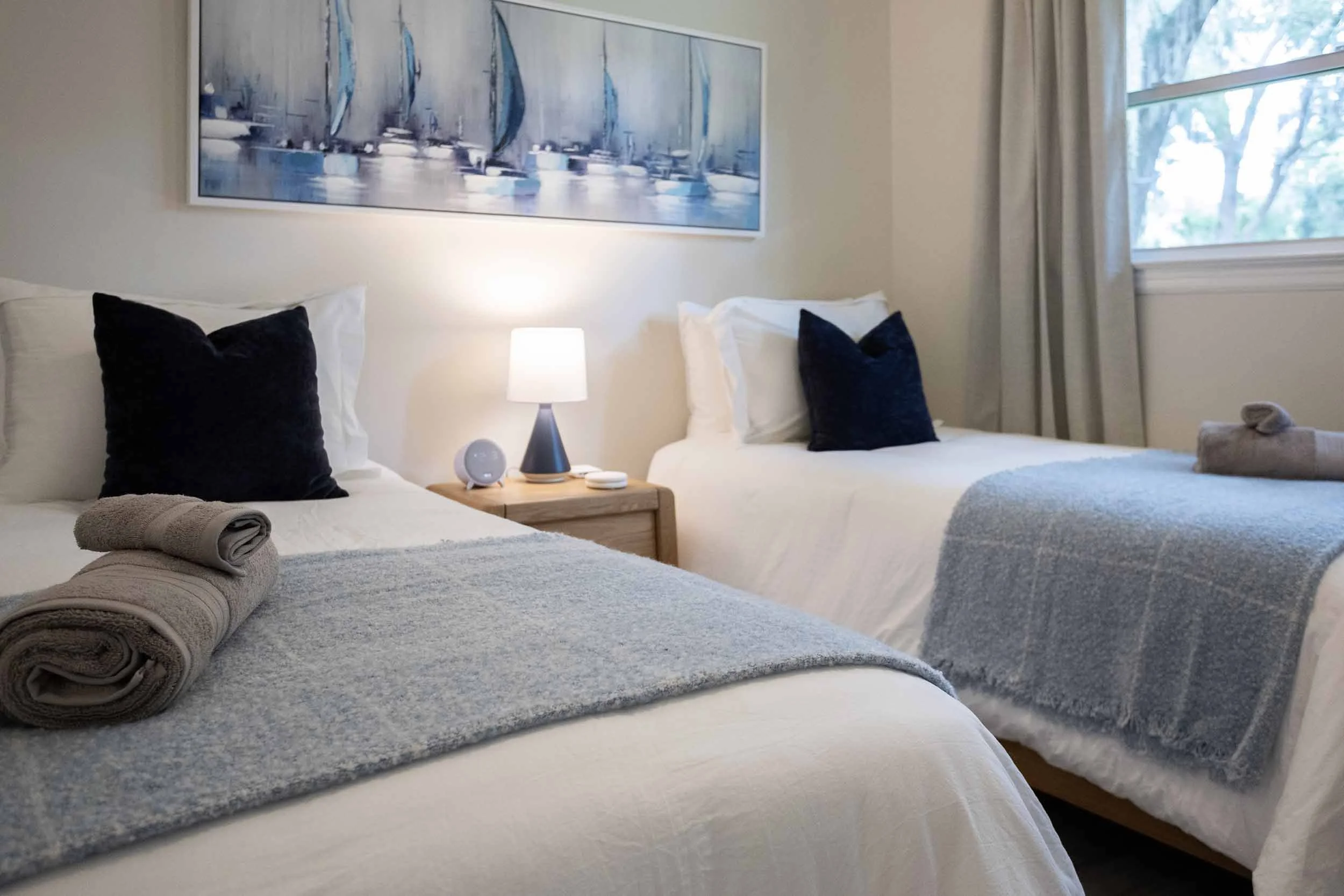 Detail view of bedroom with two twin beds, perfect for families with children, blackout curtains, pillows, throw blankets, towels, sailboat artwork, nightstand, lamp, smart speaker, charging station