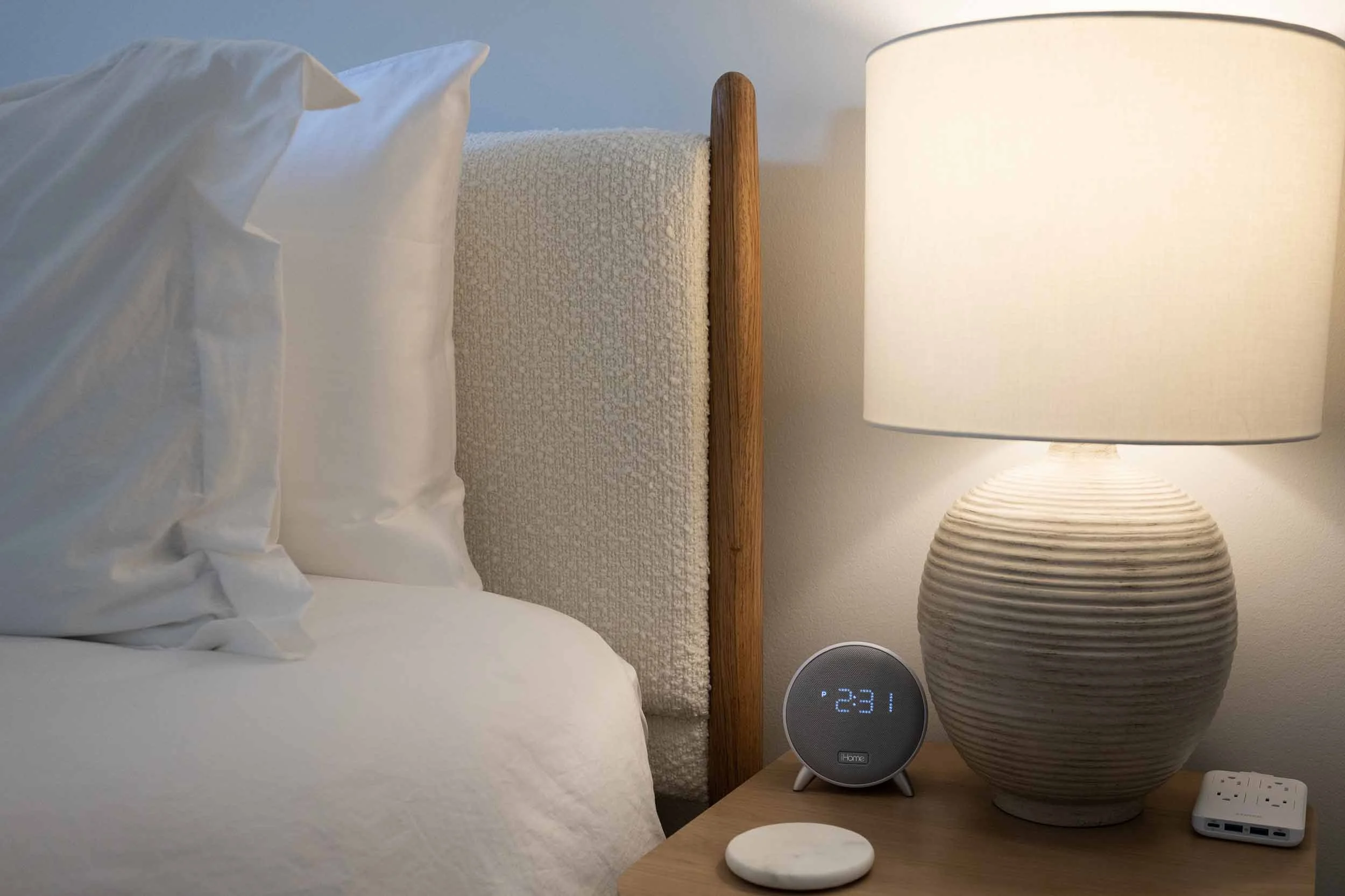 Bedside table detail of master bedroom at Pomelo House on Wilmington Island, ribbed ceramic lamp, smart speaker clock, charging station, alabster coaster, plush bedding against bouclé headboard