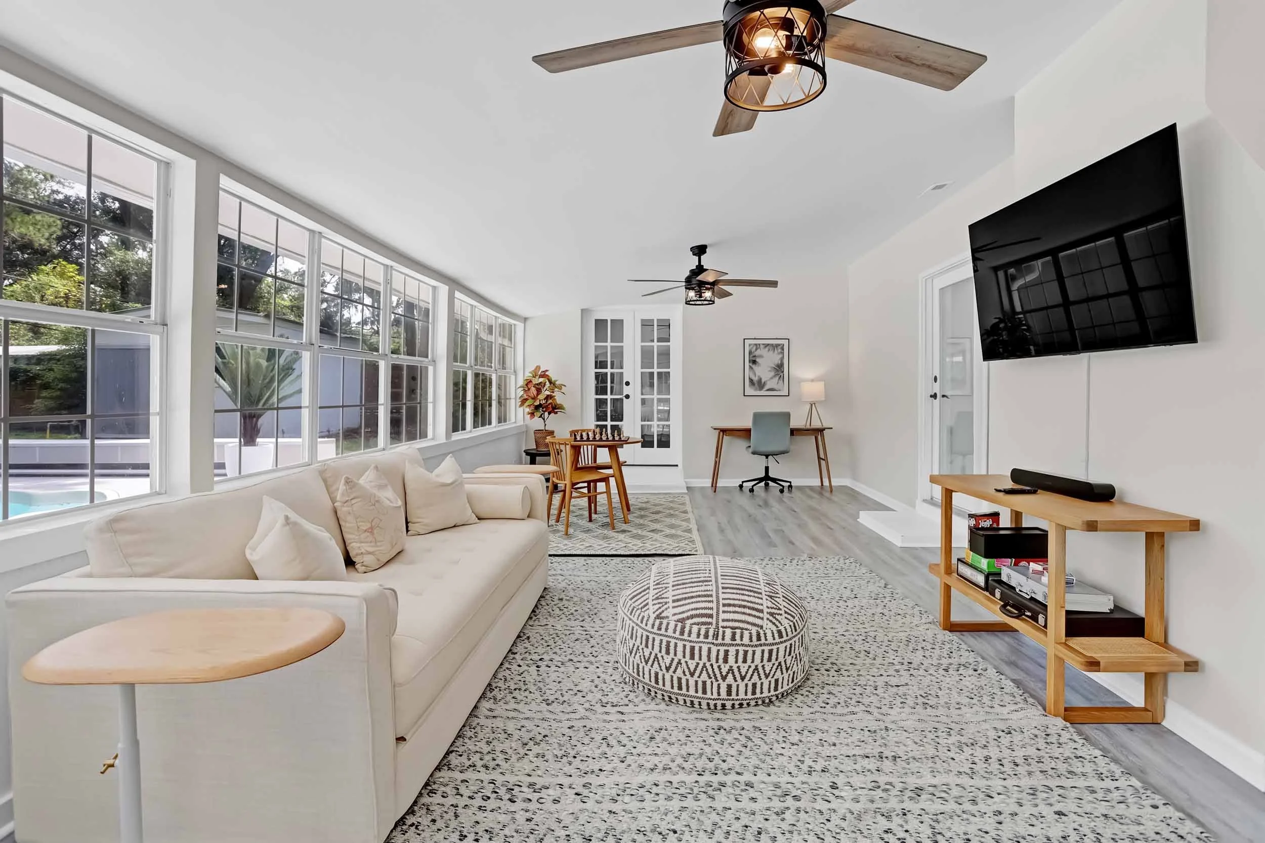 Sunroom with wall-to-wall windows overlooking pool oasis, firm sofa bed, 55” TV, console table with soundbar and board games, café table with chairs, desk with office chair, c-tables, ceiling fans