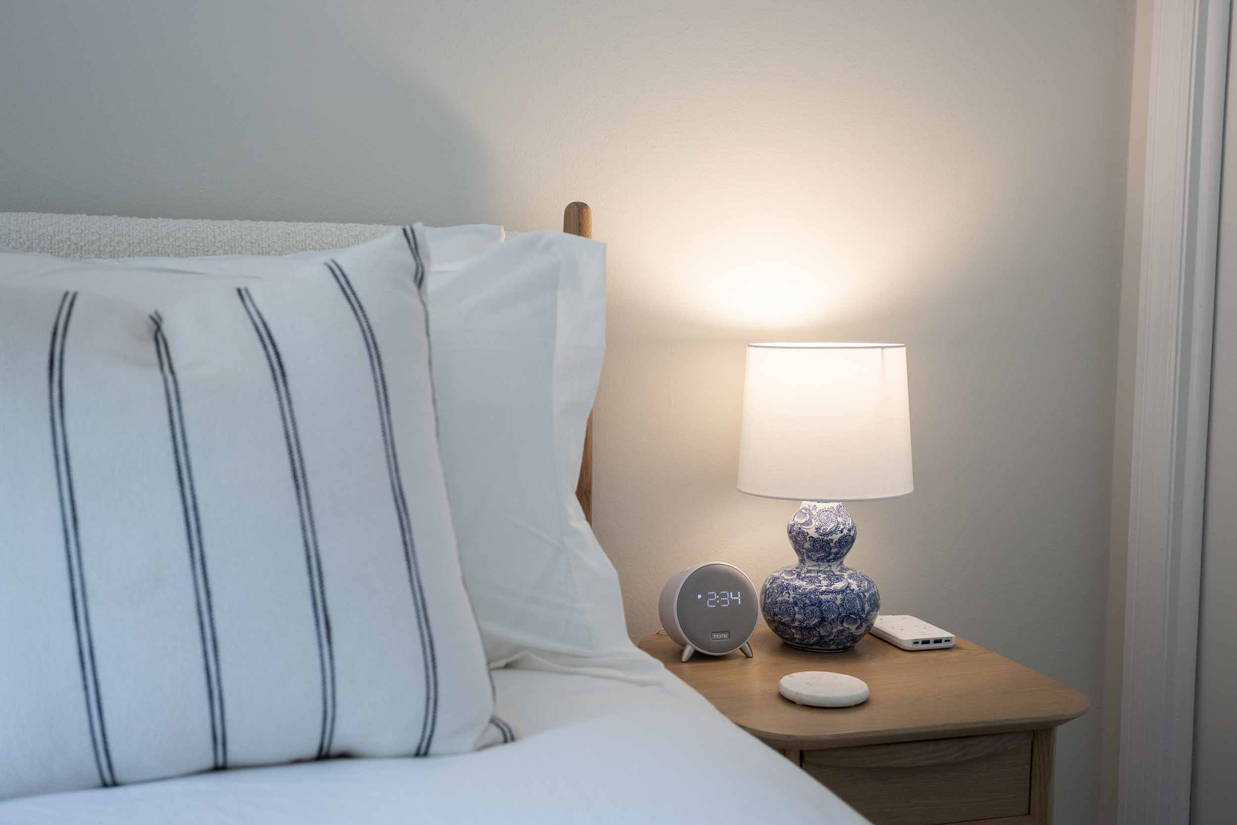 Bedside detail in hall queen bedroom, luxurious bedding with bouclé headboard, striped pillows, blue-and-white coastal lamp on white oak nightstand with smart speaker, charging station, warm lighting
