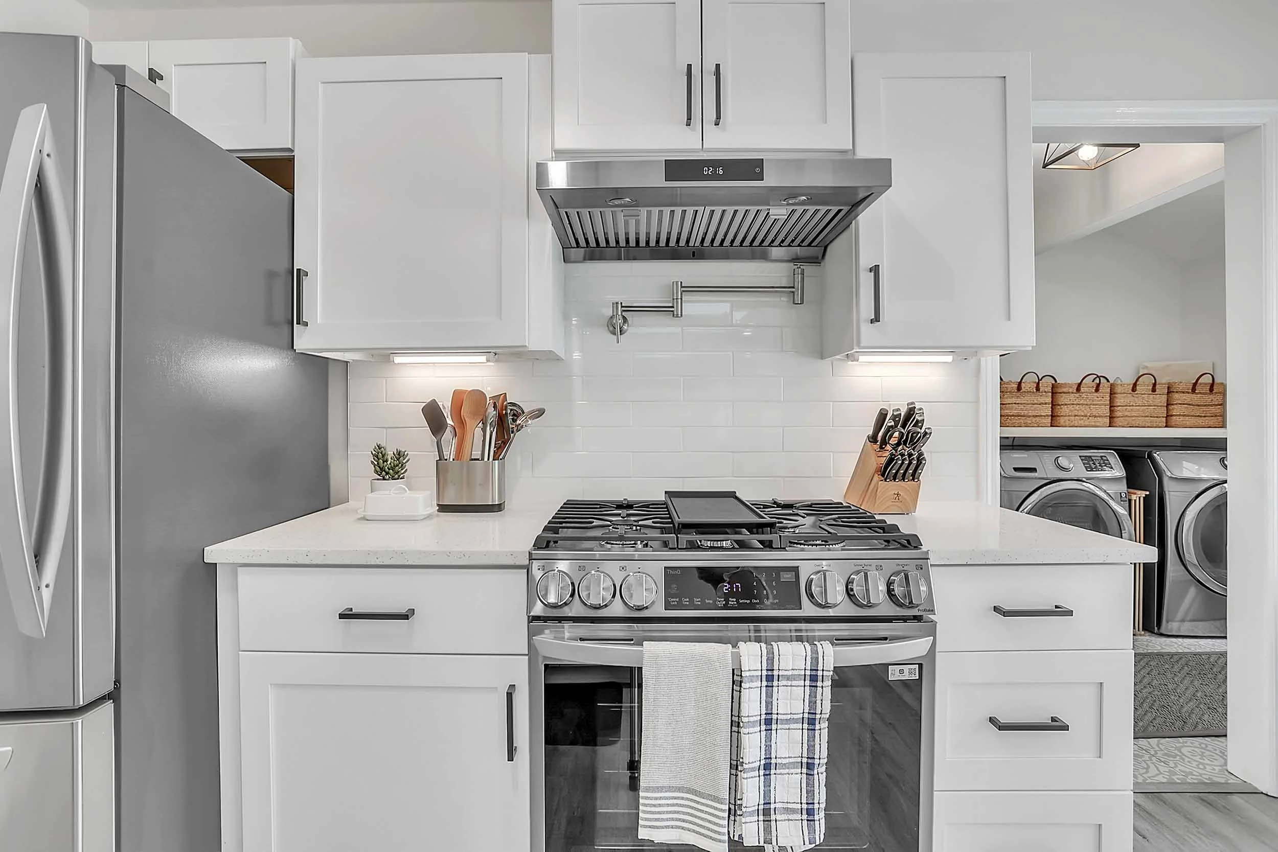 Chef’s kitchen with stainless oven and gas range with hood, white subway tiles, pot filler, refrigerator, white shaker cabinets, warm under-cabinet lighting, quartz countertops, knife block, utensils