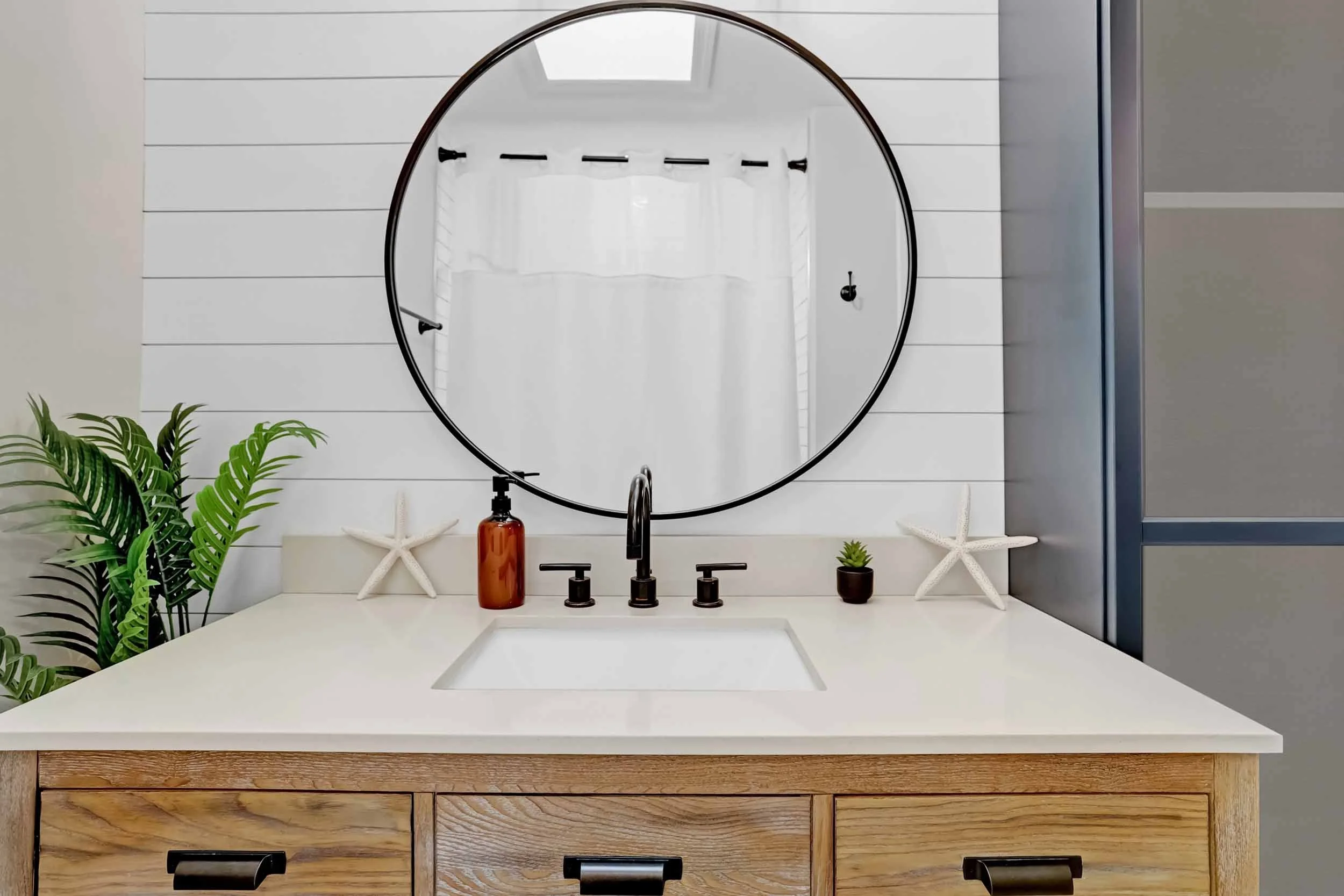 Closeup of beach-themed hall bathroom showing wood vanity with sink and sand-colored countertop, bronze faucet, round mirror, shiplap wall, tropical palm plant, starfish decor, succulent, hand soap