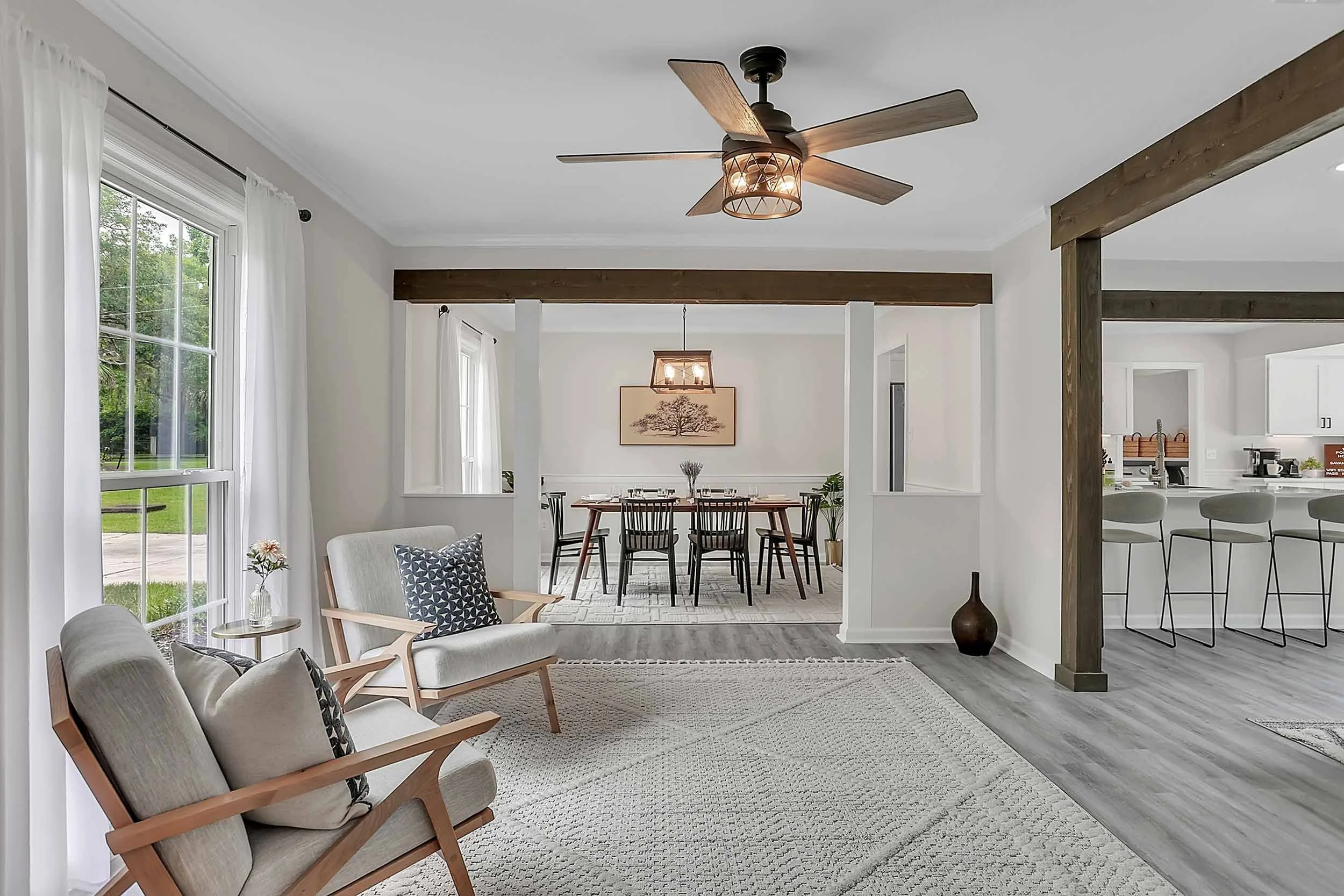 Airy coastal sitting room with mid-century modern lounge chairs, throw pillows, and side table with marble base by oversized window, white sheer curtains, trellis area rug, wood beams, ceiling fan