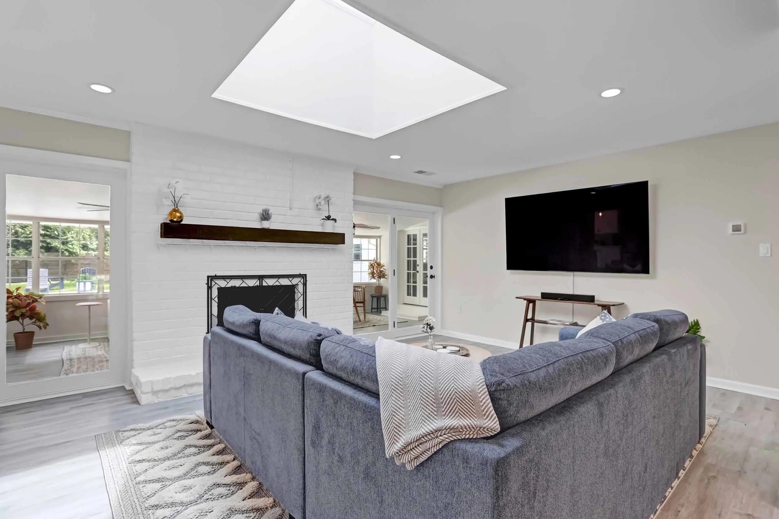 Open living room with large skylight, blue sectional with throw pillows and blanket, white brick fireplace with mantel, 77” TV, console table with soundbar, french doors to sunroom, view of pool oasis