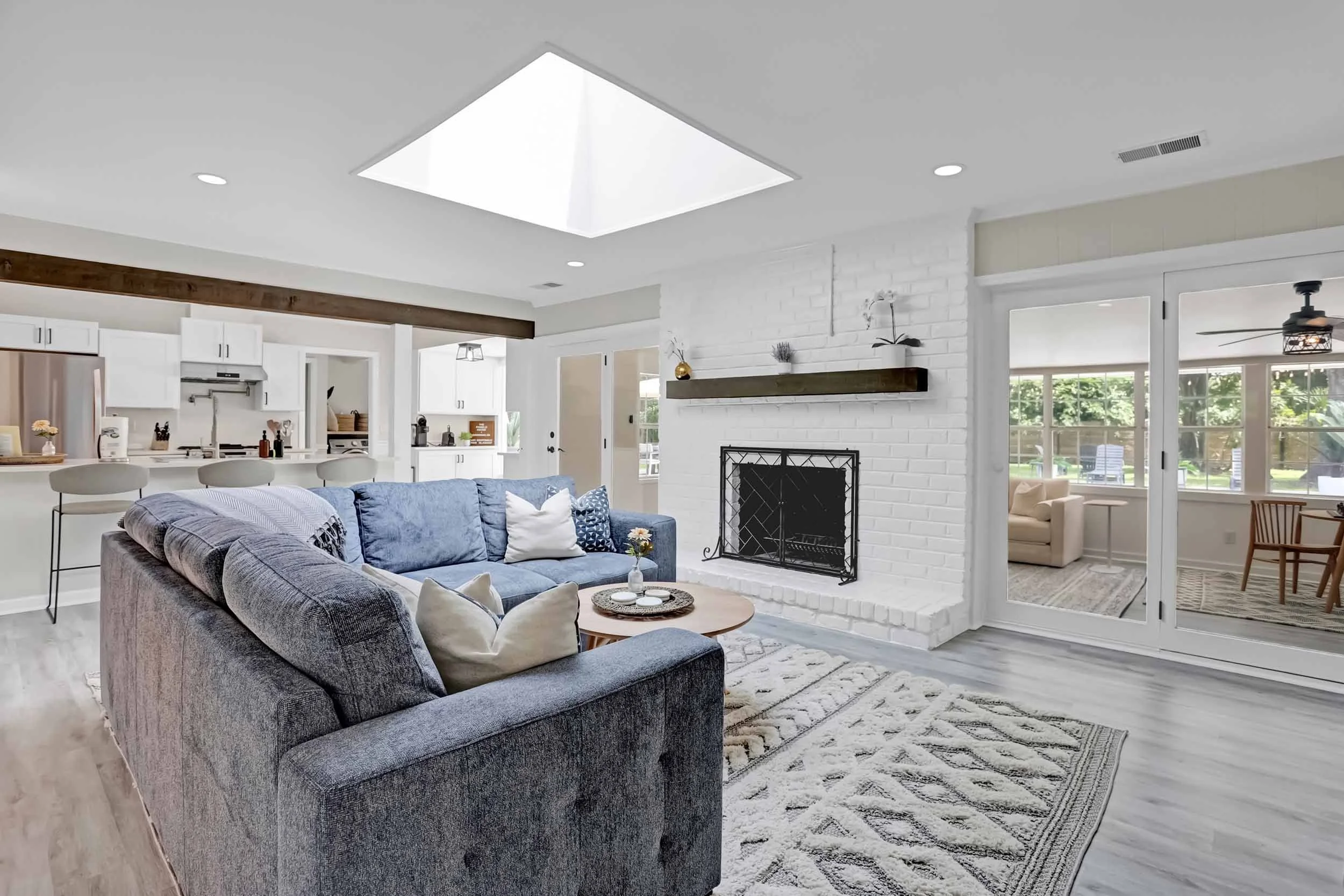 Alternate view of living room showing white brick fireplace, wood mantel, oversized skylight, view of chef’s kitchen, multiple french doors opening to sunroom, with clear view of private heated pool