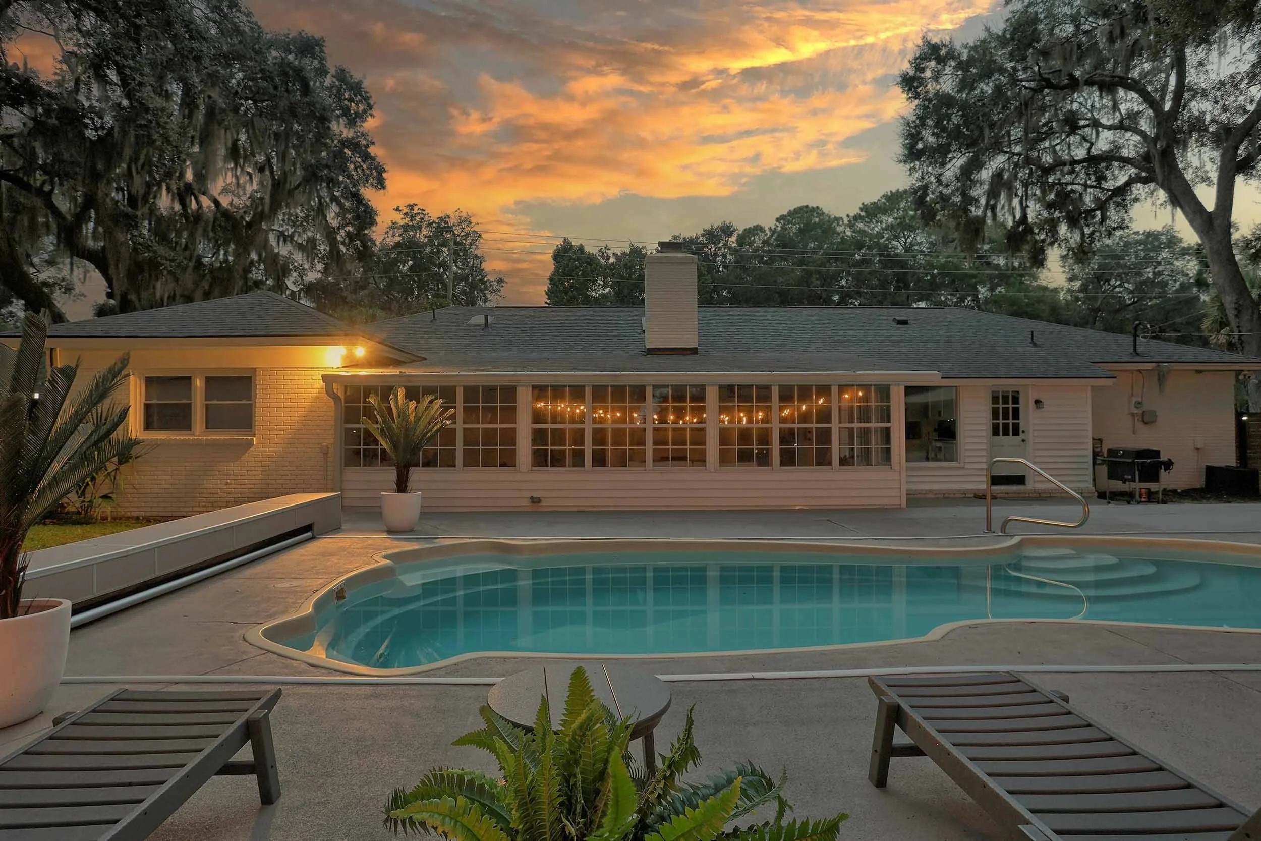 Sunset view from chaise lounges by heated pool in private backyard oasis, between Tybee Island and Savannah’s historic downtown, with warm reflection of glowing string lights in sunroom windows