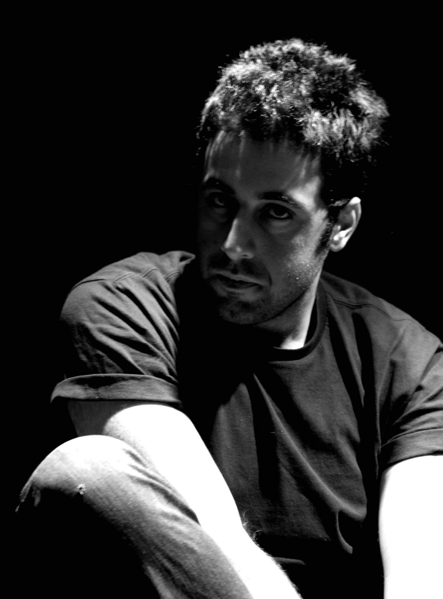 Black and white photo of a young man with curly hair, wearing a sweatshirt, sitting against a solid black background, with a serious expression on his face.