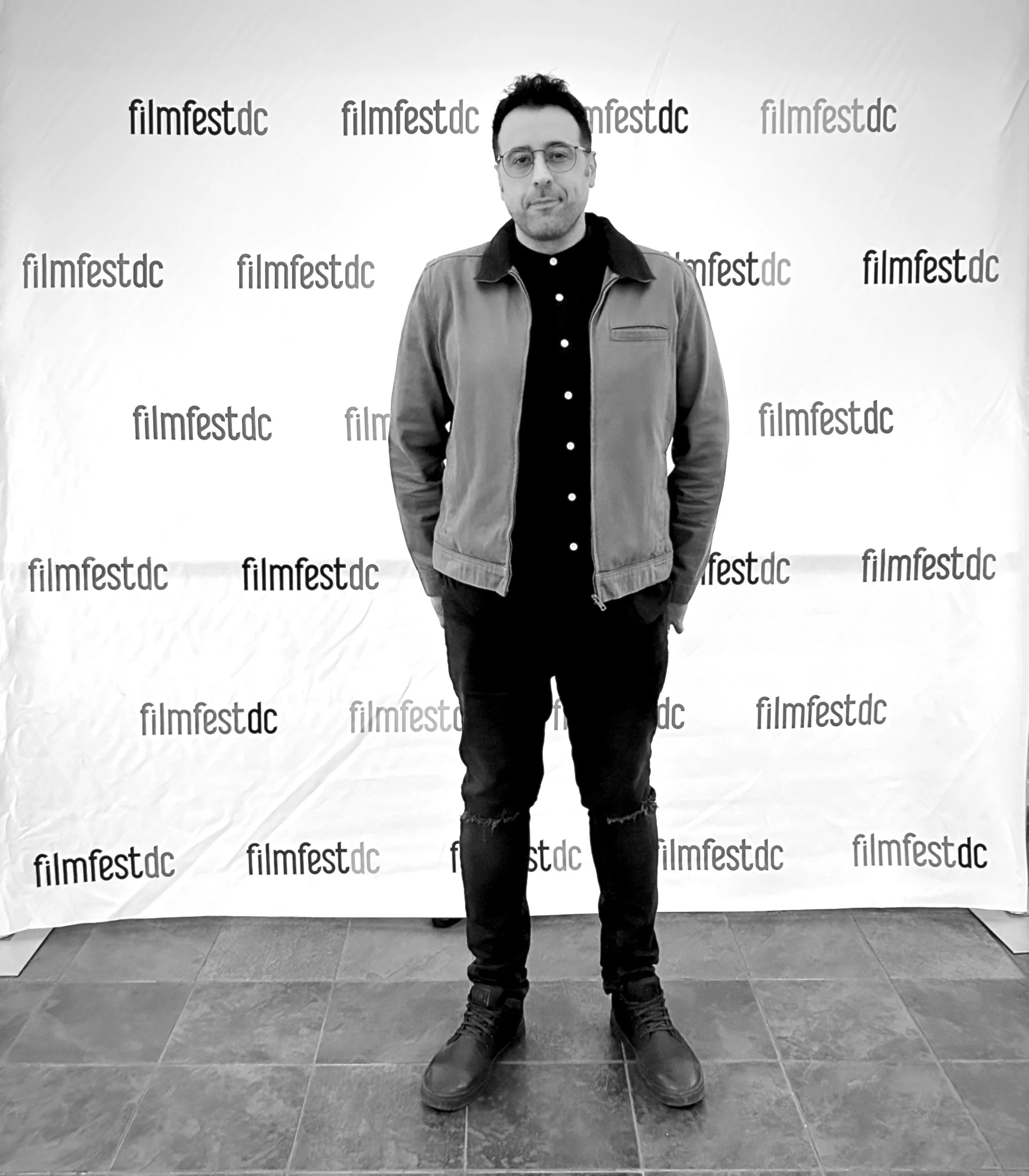 Man wearing glasses, black shirt, and light jacket, standing in front of a step and repeat backdrop with 'filmfestdc' repeated on it, in an indoor setting with gray tile floor.