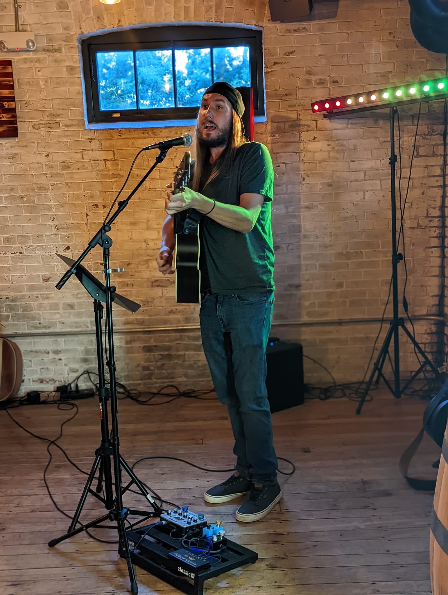 A man with long hair and a dark beanie playing an acoustic guitar and singing into a microphone in a cozy brick-walled venue with a small window and colorful lighting.