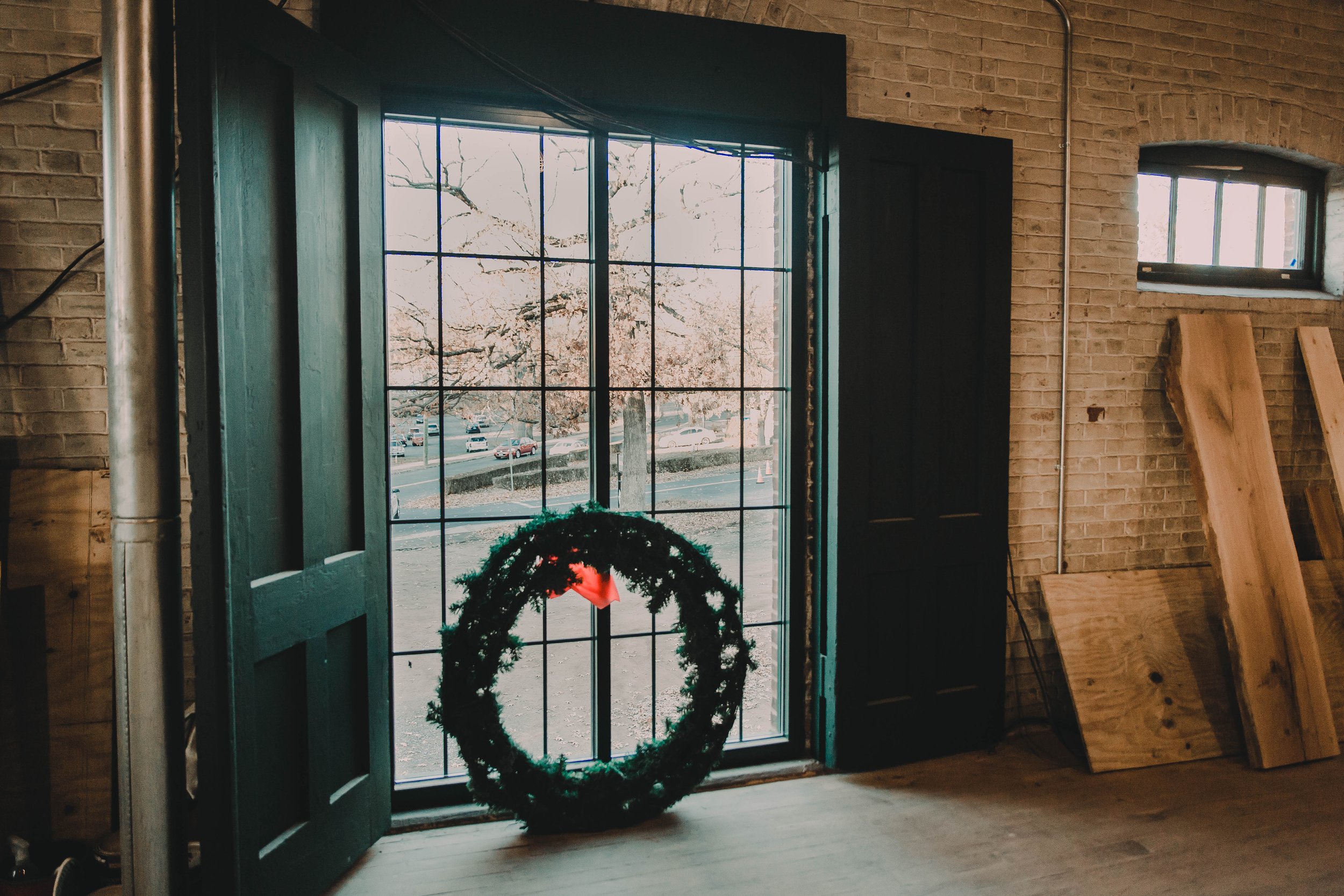Interior of a room with large open window and a decorative wreath leaning against window frame, with wooden planks stored on the floor and a brick wall in the background.