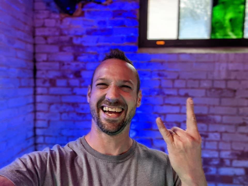 A man with a beard smiling and making a rock on hand gesture, standing in front of a brick wall with colorful lighting.