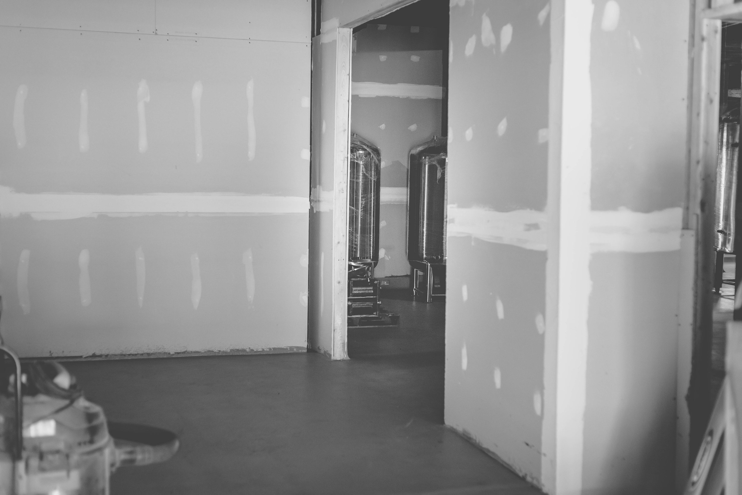 Black and white photo of an unfinished room with drywall and two water tanks visible in the background.