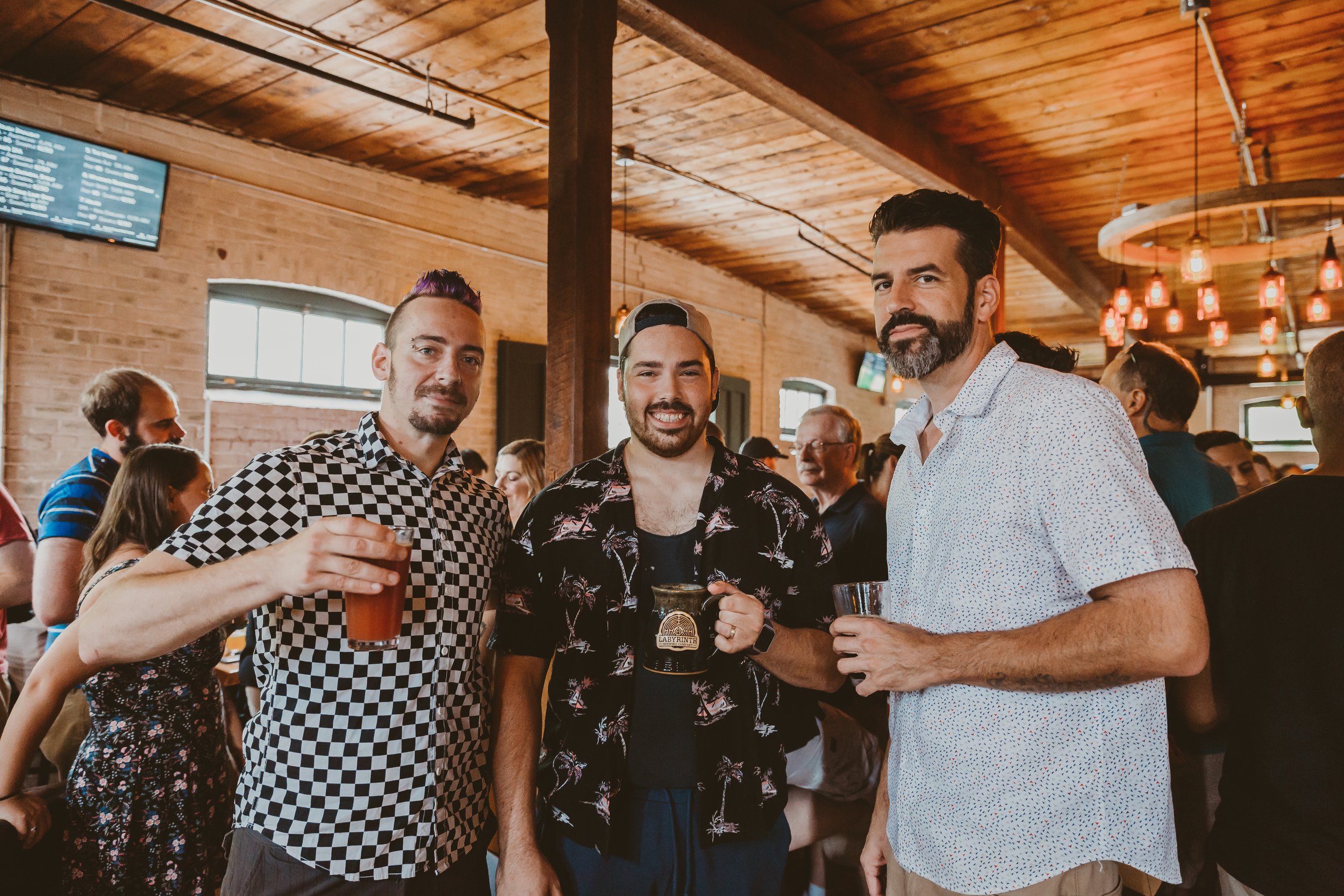 Three men are standing together in a crowded bar or brewery, holding drinks and smiling at the camera. The setting has warm lighting, wooden ceiling, brick walls, and large windows.