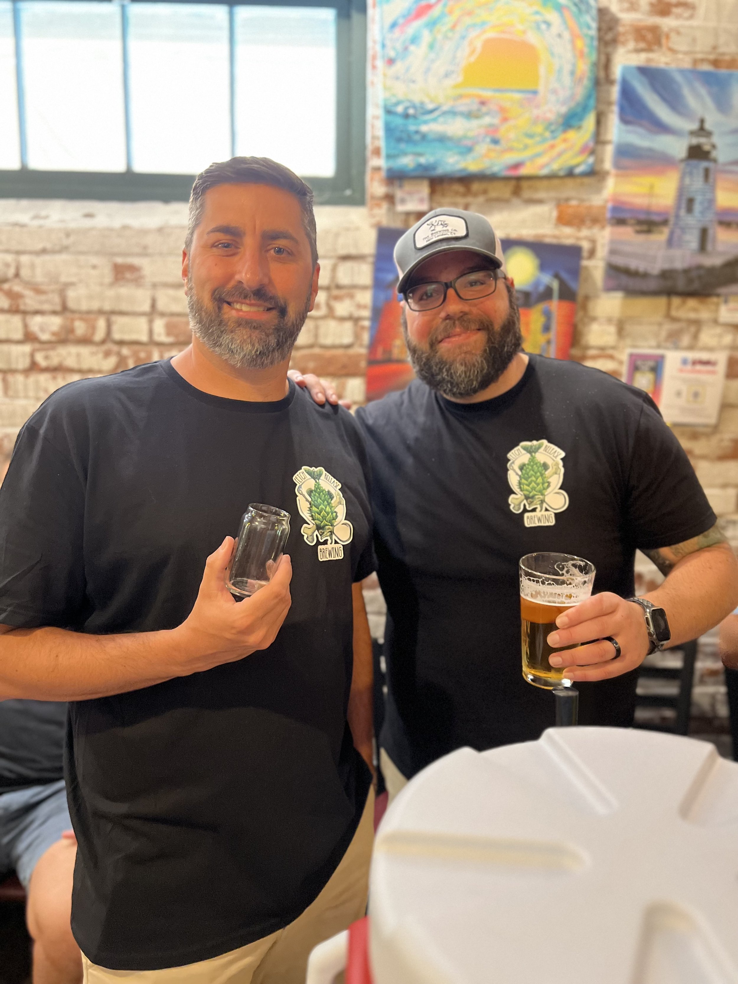 Two men smiling indoors, holding drink glasses, wearing matching black t-shirts with a green hop logo, with colorful paintings and a brick wall in the background.