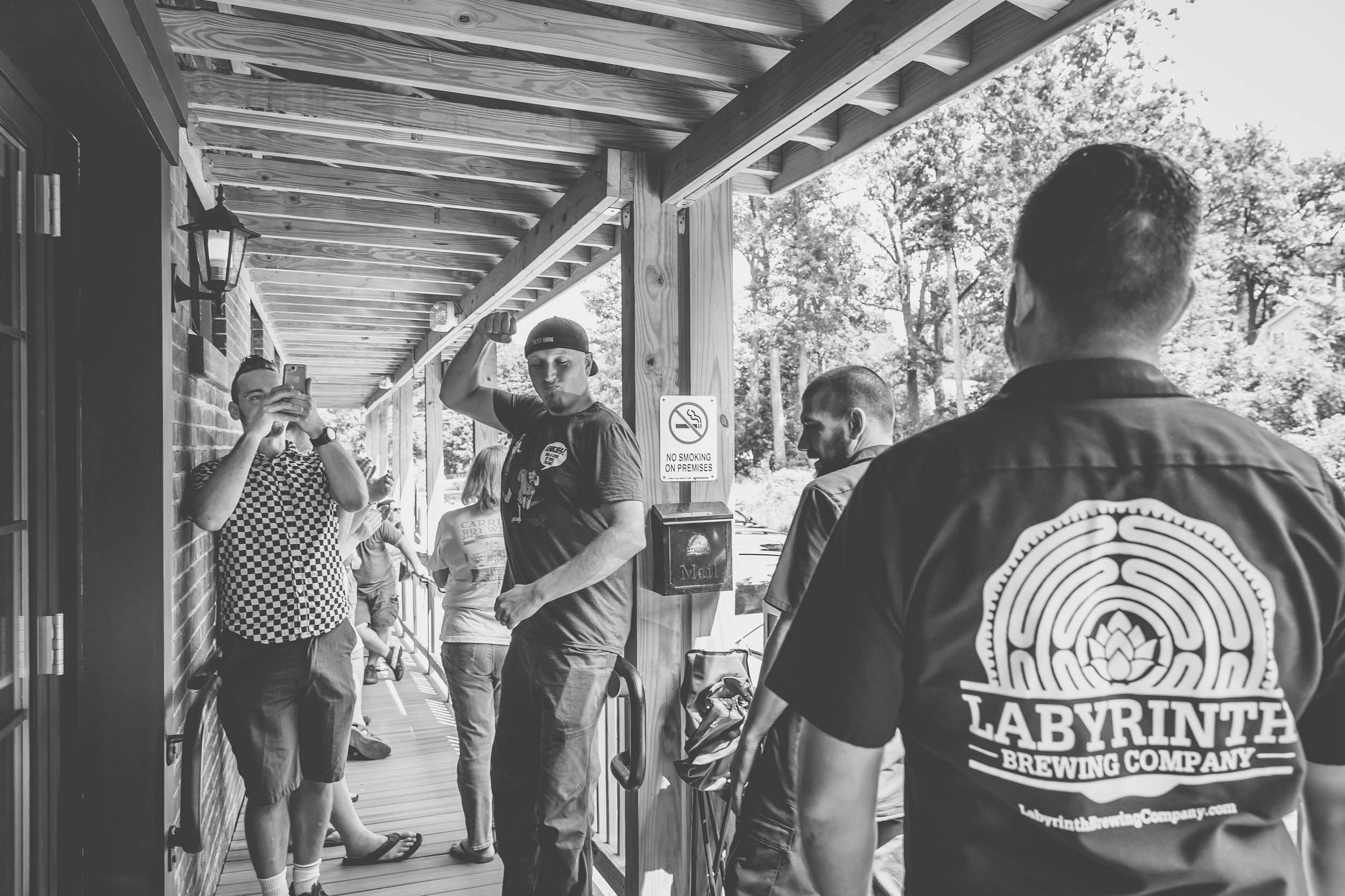 Group of people on a porch, one man taking a photo with his phone, others talking, and a man wearing a Labyrinth Brewing Company shirt, outdoors with trees in the background, black and white photo.