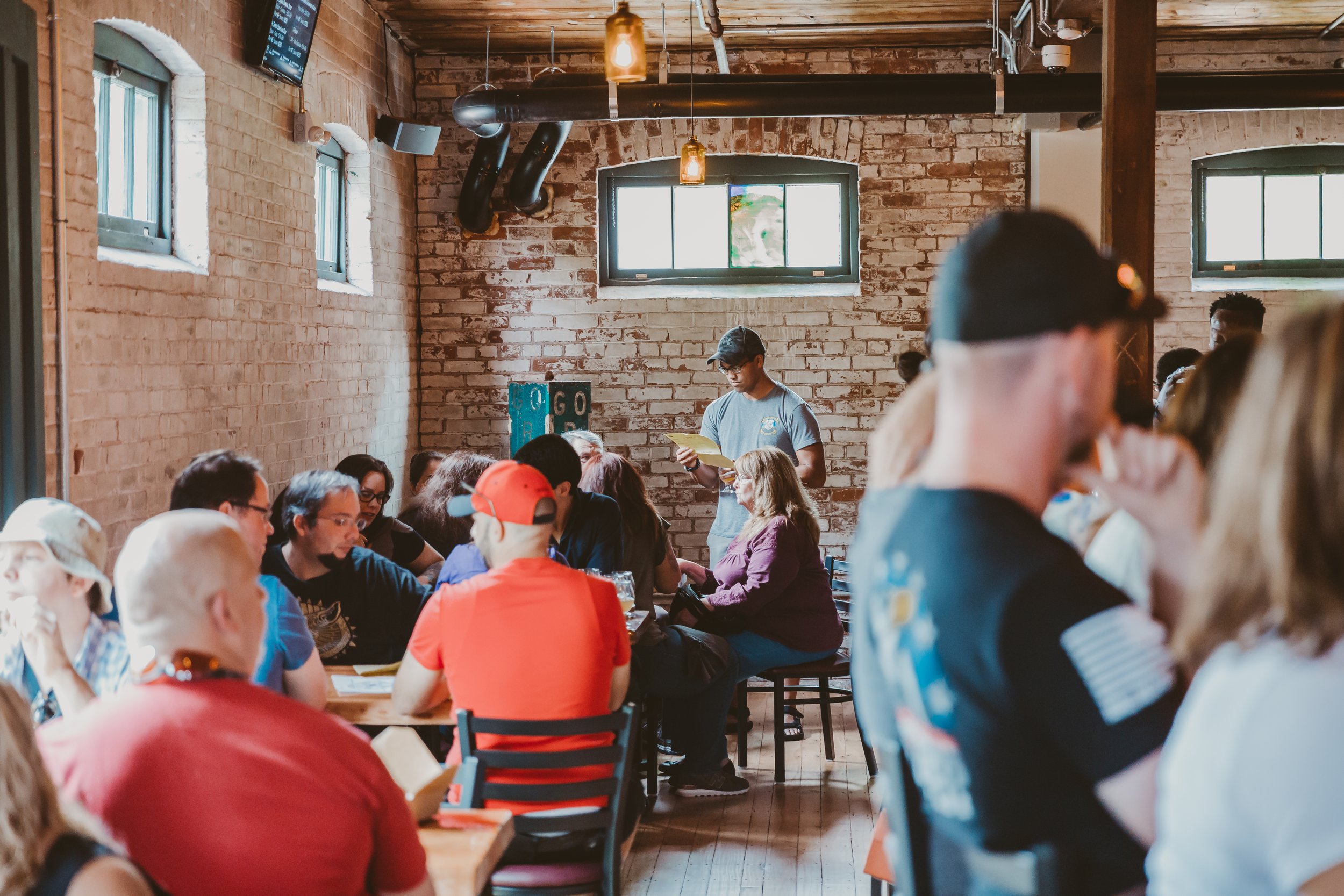 People gathered in a brewery or cafe with exposed brick walls, some reading or talking, while a person stands at a table reading aloud to the group.